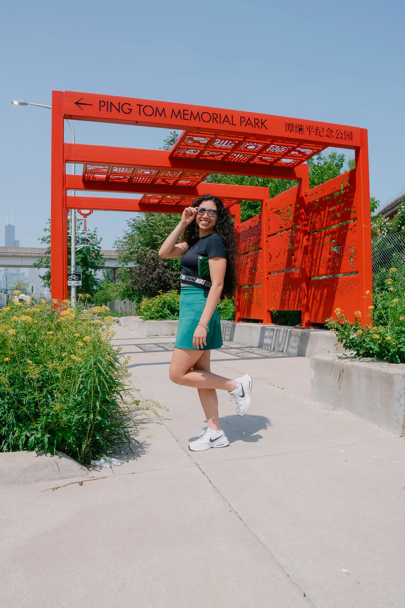 A woman with curly hair wearing sunglasses, a black t-shirt, teal skirt, and white sneakers smiling and posing on a sidewalk near a bright orange structure that reads "Ping Tom Memorial Park" in both English and Chinese. Green foliage and a city skyl