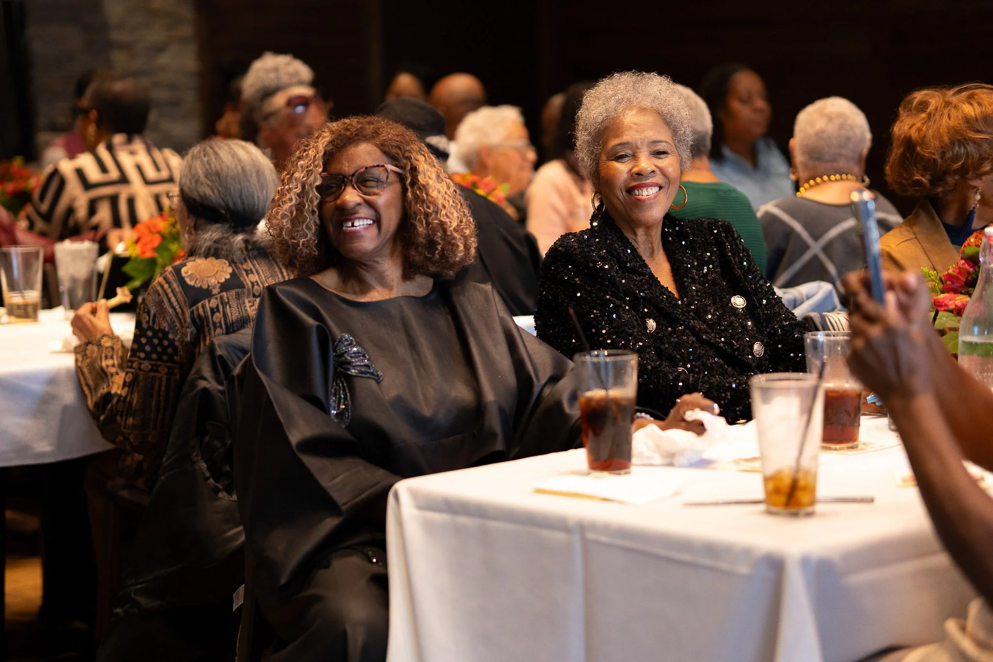 Two smiling women sitting at a table at a social event, surrounded by other guests and floral arrangements.