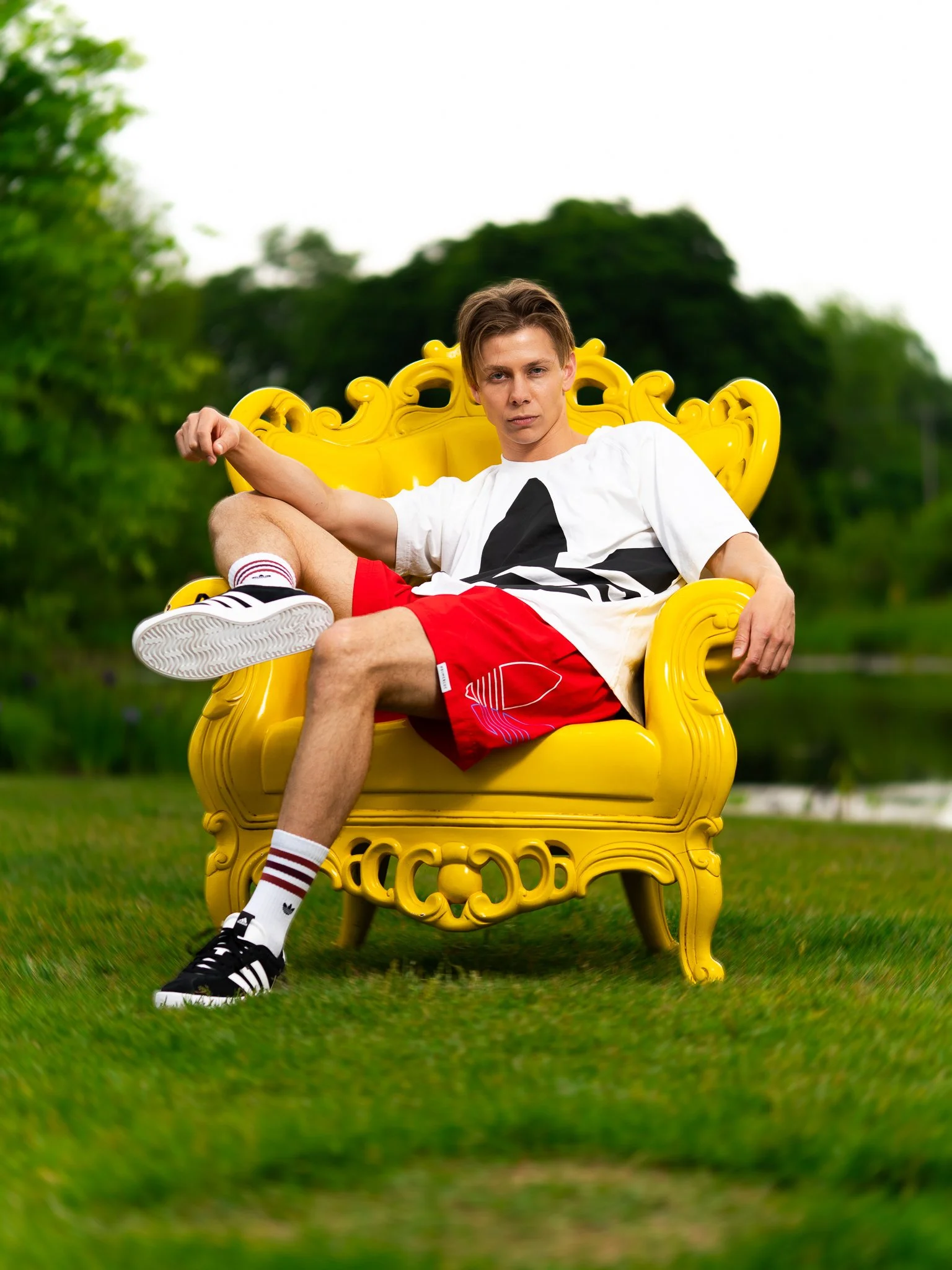 Young man sitting on an ornate yellow outdoor chair on grass, wearing a white T-shirt, red shorts, and black sneakers.