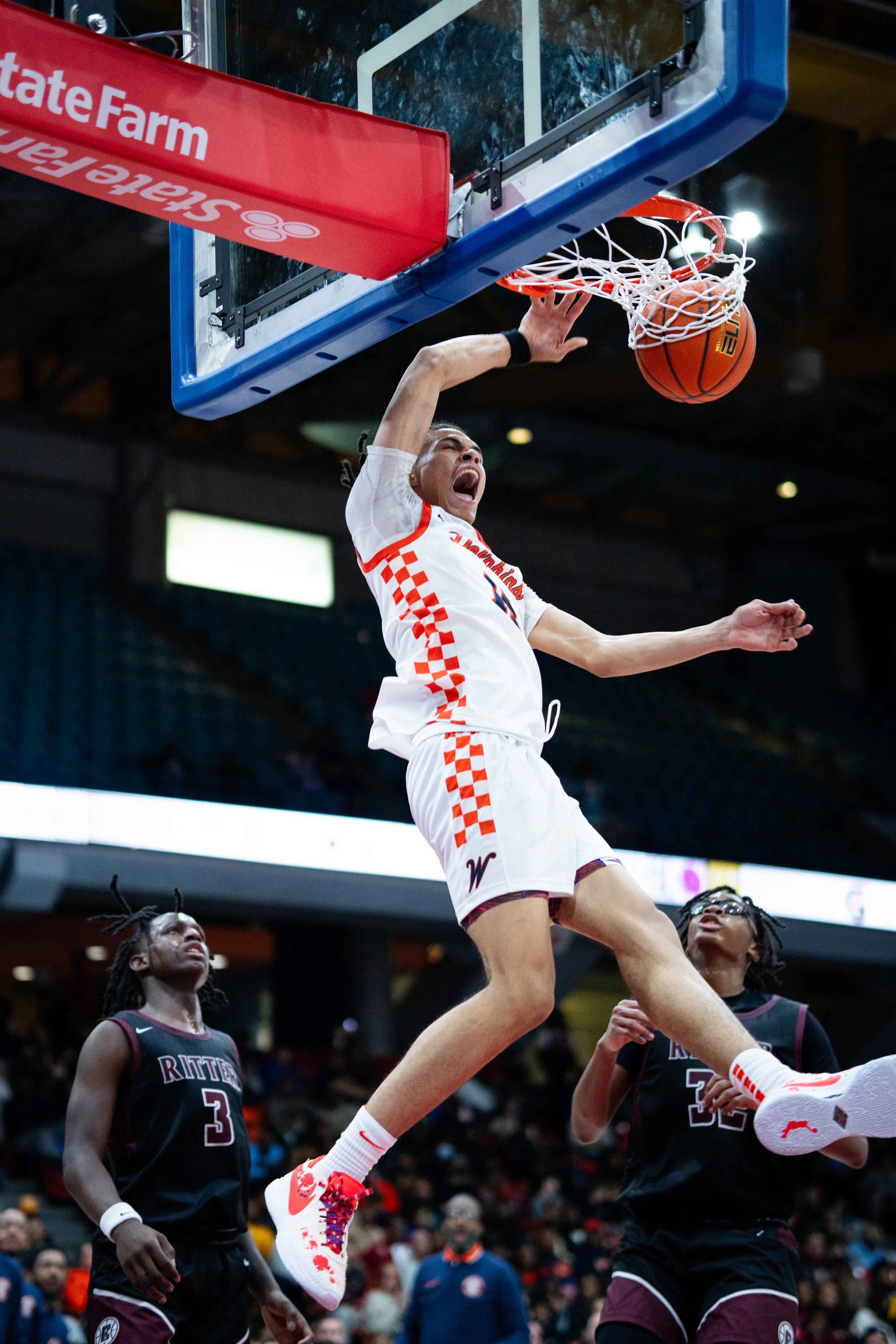 A basketball player in a white and orange checkered uniform dunking the ball into the hoop while two players in black uniforms observe from below.