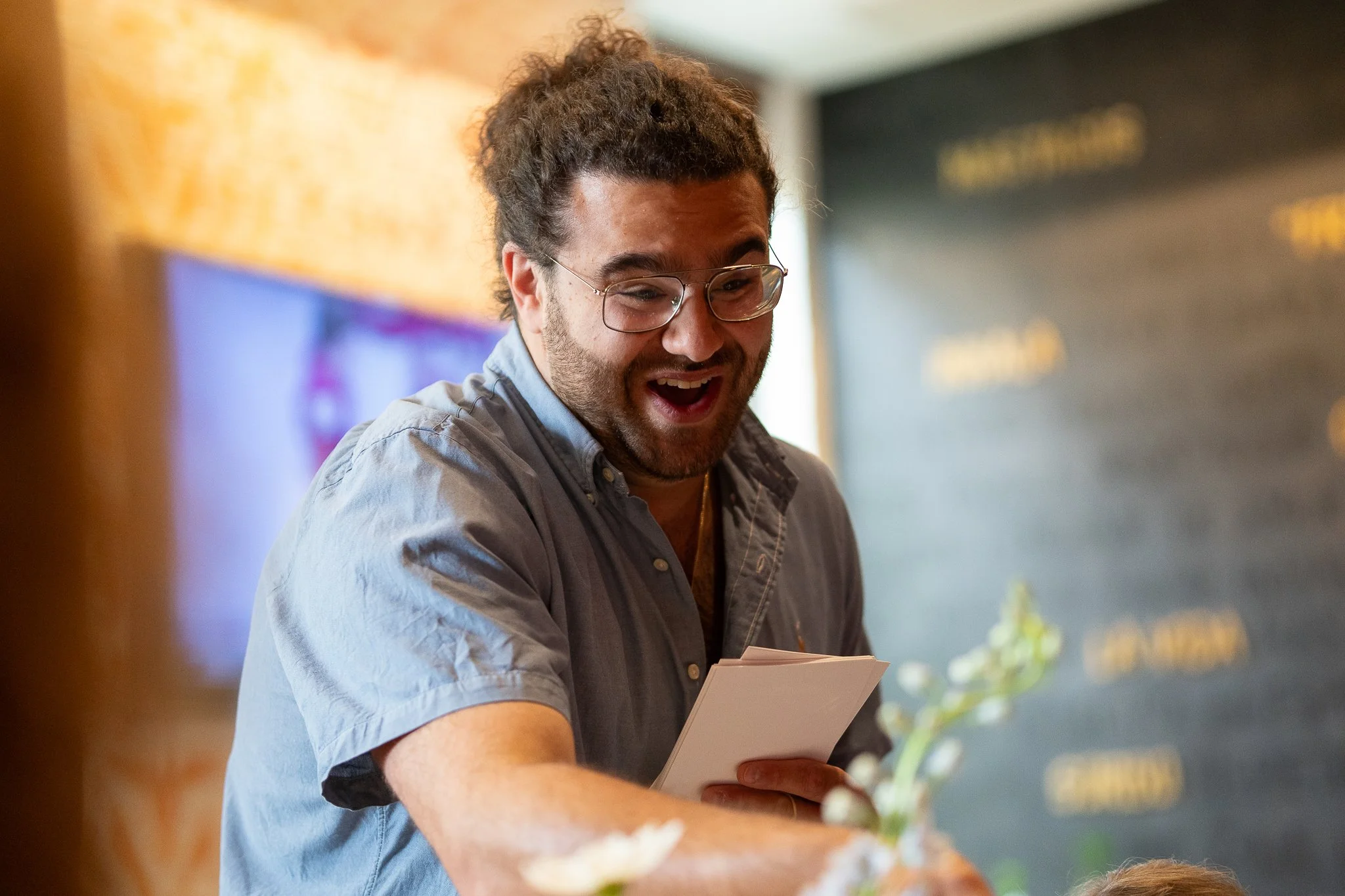 A man with glasses and curly hair, wearing a gray button-up shirt, smiling and holding papers, in a casual indoor setting.