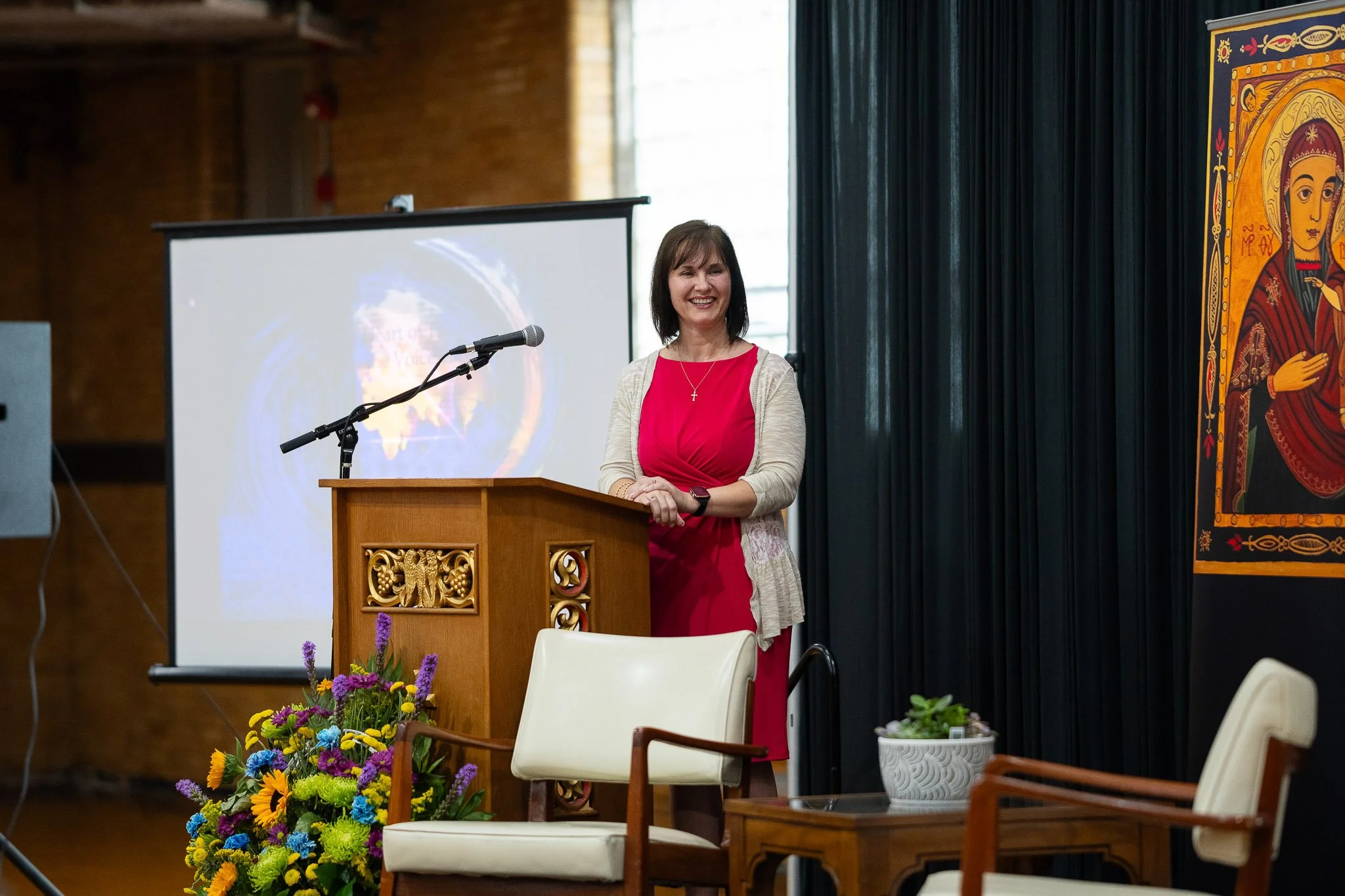 A woman in a red dress and beige cardigan stands at a wooden podium in a room with dark curtains, a large screen behind her, and a colorful religious icon on the wall. There are chairs, a potted plant, and a bouquet of flowers in front of her.