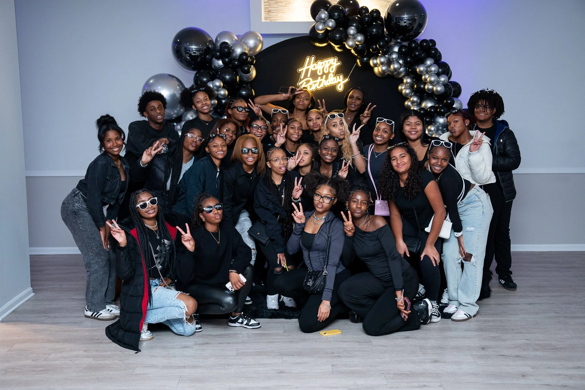 Group of young women and girls celebrating a birthday in front of a black and gold balloon backdrop with a neon 'Happy Birthday' sign, posing and smiling for the photo.