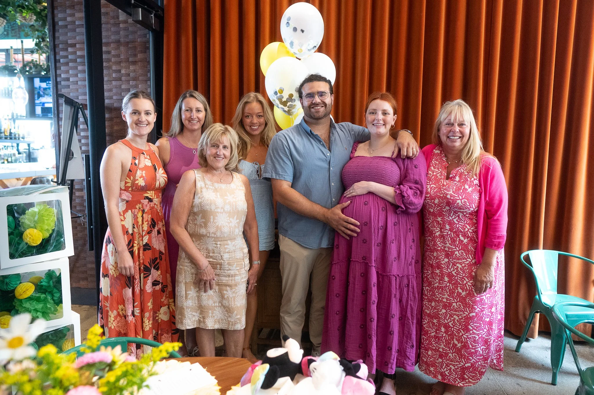 Group of eight women and one pregnant woman in a pink dress during a baby shower, standing in front of a brown curtain with balloons in the background.