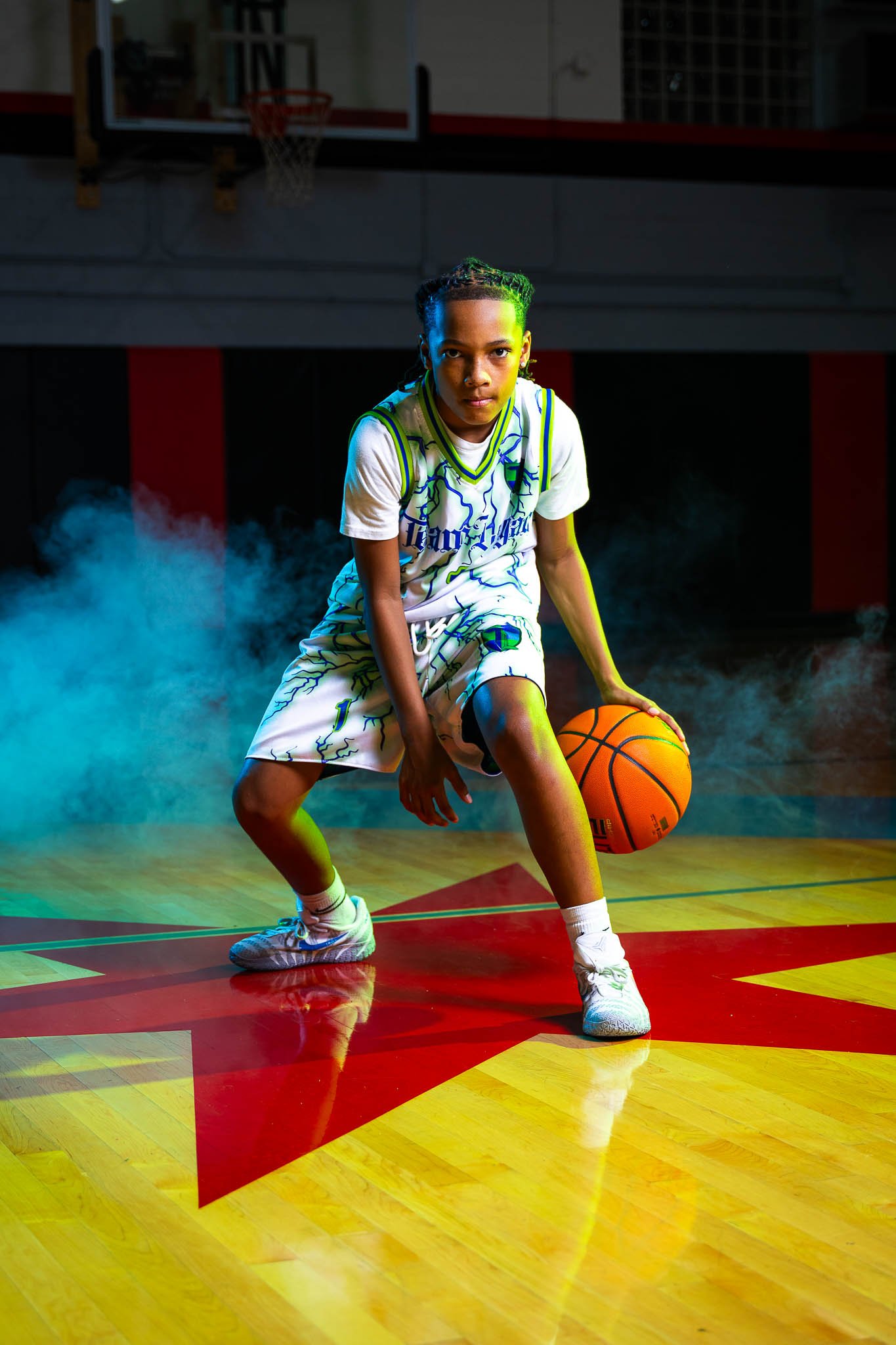 A young boy in a basketball uniform crouches with a basketball in his right hand on a basketball court with smoke effects.