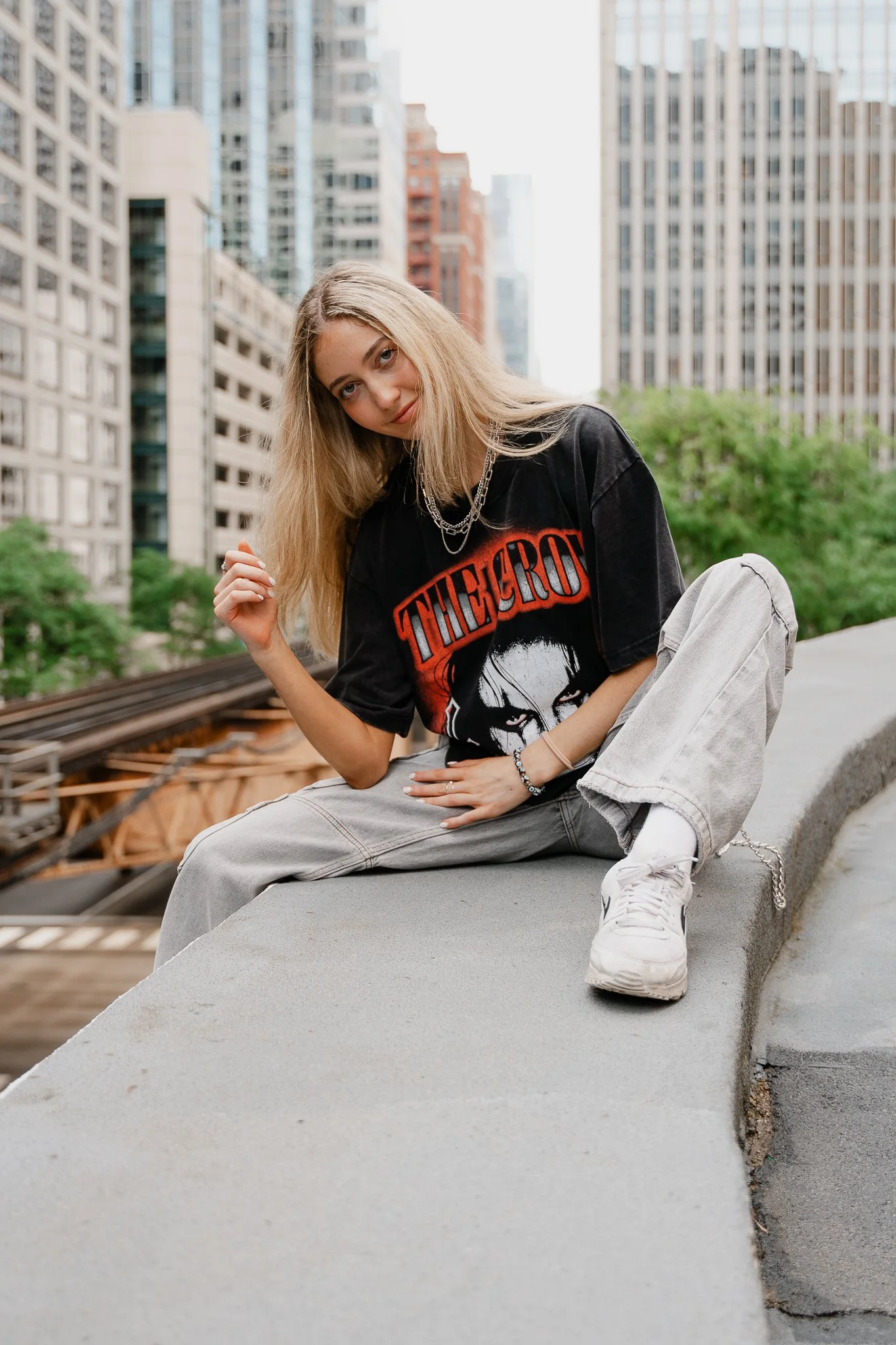 A young woman with blonde hair wearing a Black Sabbath t-shirt, light grey jeans, and white sneakers, sitting on a concrete ledge in an urban setting with tall buildings and trees in the background.