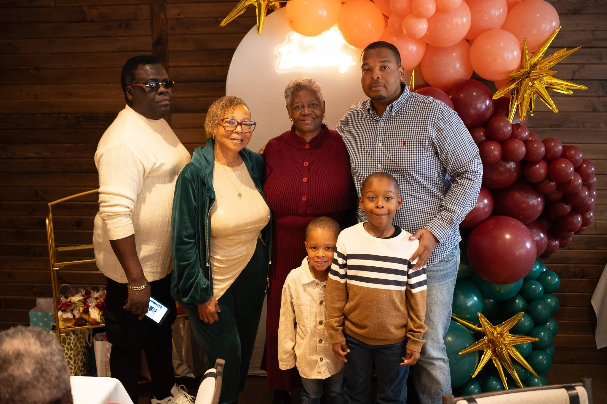 A family portrait of seven people standing in front of a balloon decoration with pink, burgundy, and teal balloons, with a wooden wall background. They are indoors, celebrating an event.