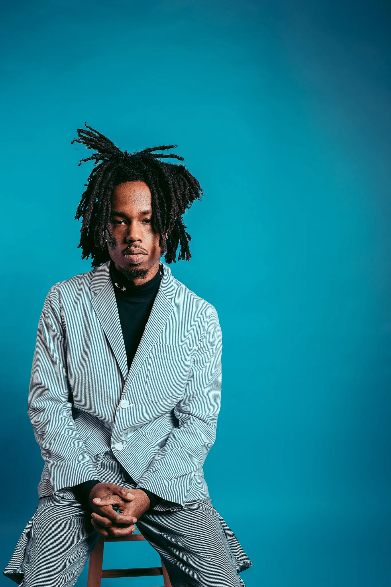 A man with dreadlocks sitting on a stool, wearing a striped blazer over a black turtleneck, against a blue background.