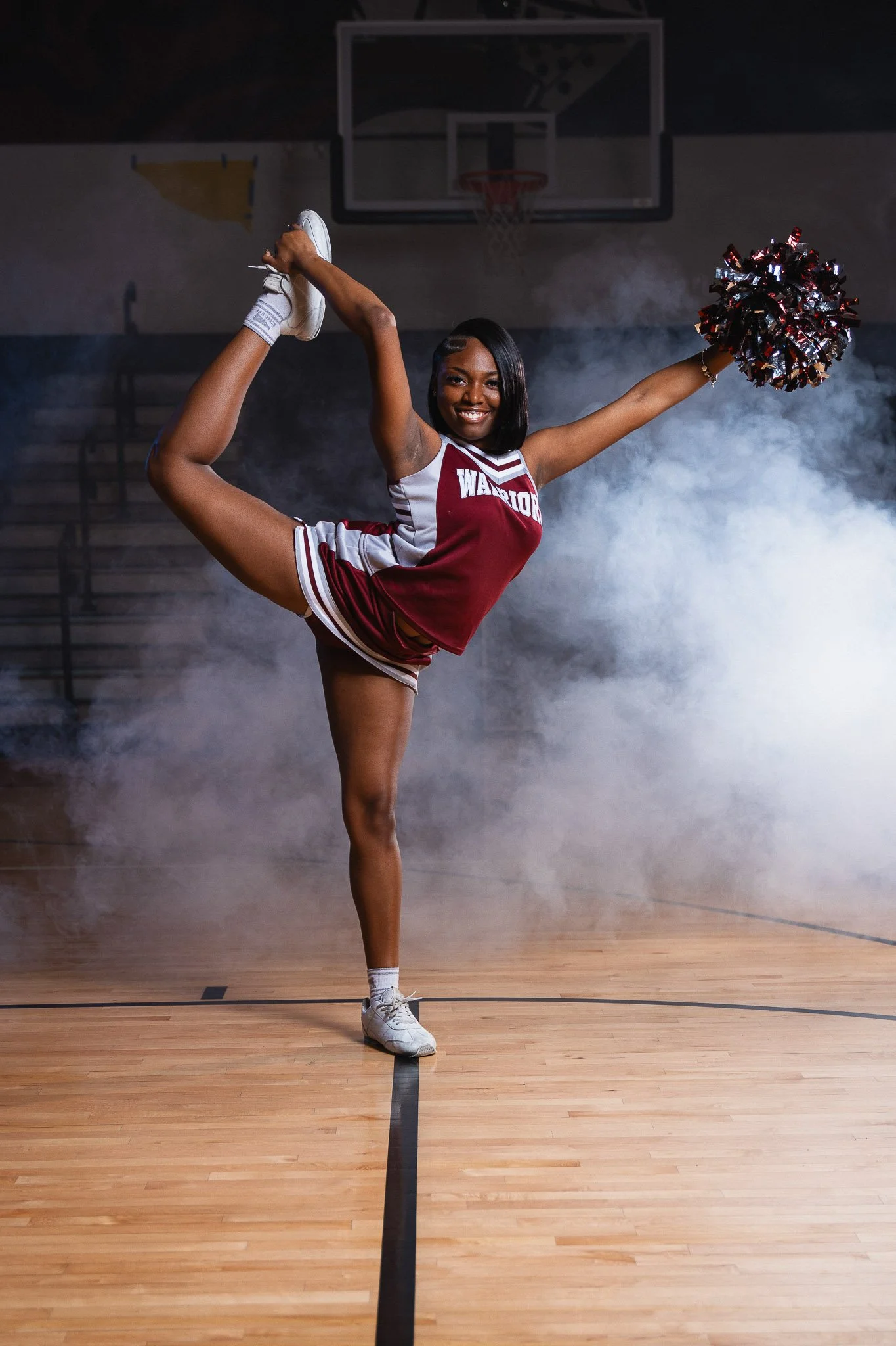 A young female cheerleader in a maroon and white uniform performs a high kick on a basketball court, holding pom-poms in one hand, with a basketball hoop and smoky background in the gym.