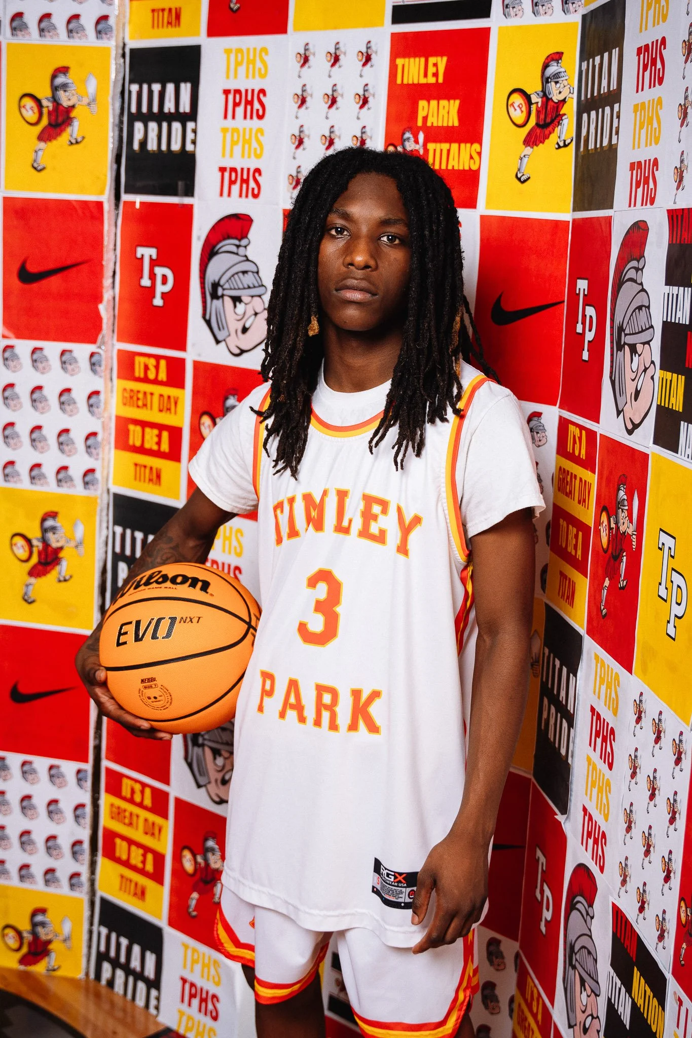 A young male basketball player standing in front of a wall decorated with school and sports logos, wearing a white basketball uniform with red and yellow accents, holding an orange basketball in his right hand.
