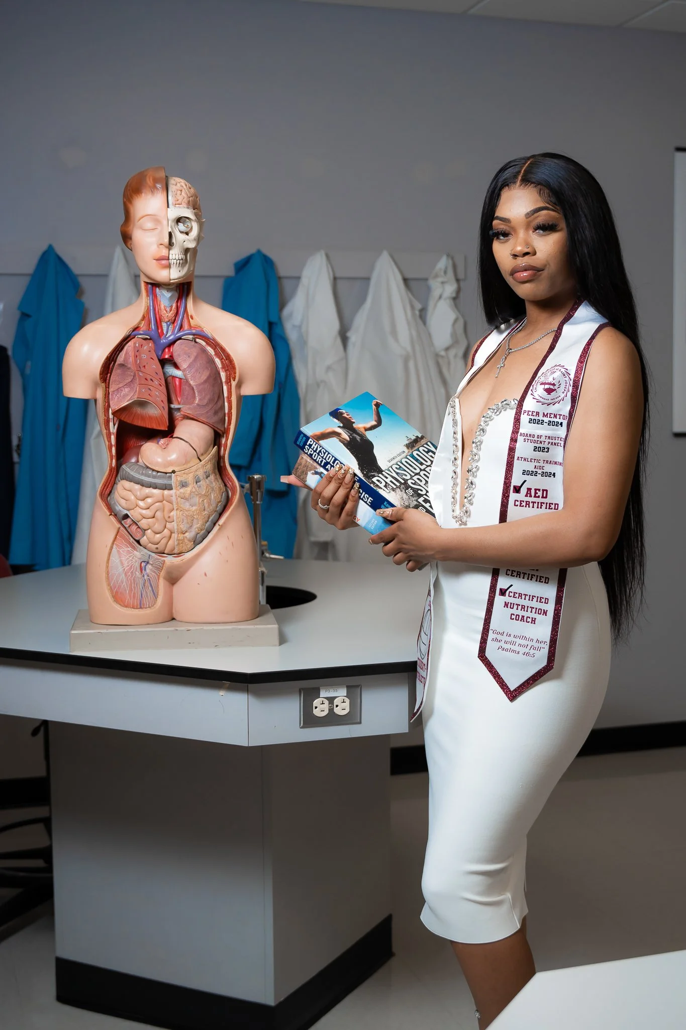 A woman in a white medical or nursing uniform standing next to a human anatomical model of the torso showing internal organs in a classroom or laboratory setting.