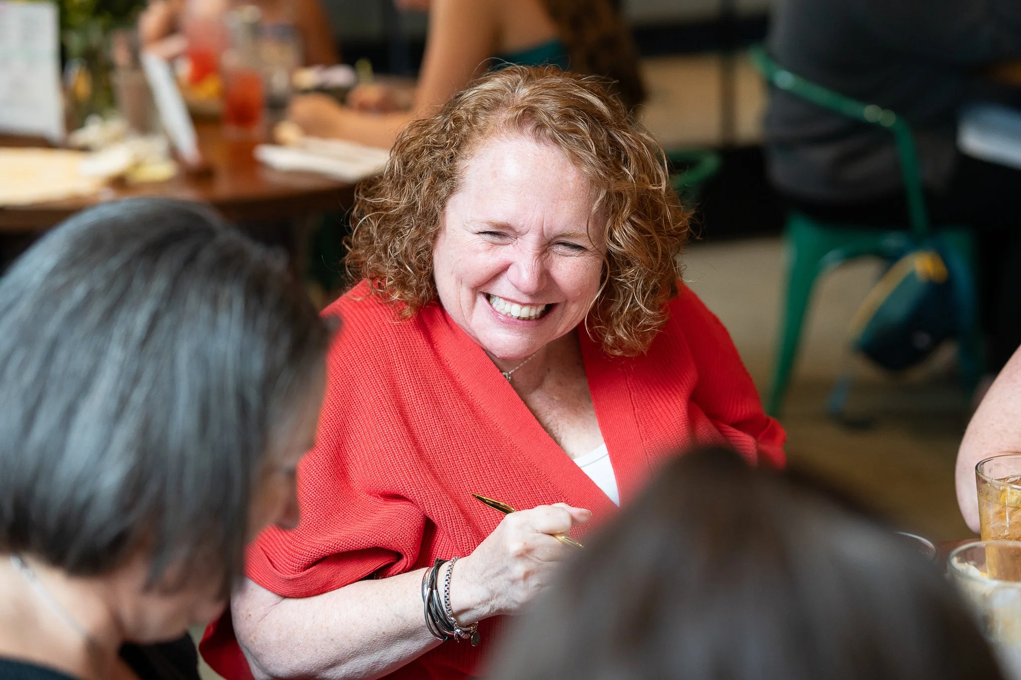A woman with curly red hair wearing a red top, smiling and talking with others at a restaurant or cafe table.