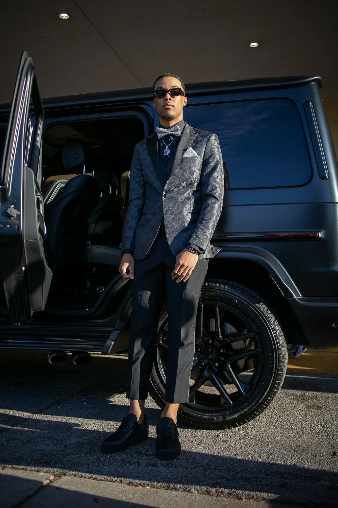 A man wearing a patterned blazer, black pants, and black shoes, standing next to an open door of a black luxury SUV, dressed in formal attire with sunglasses and a bowtie.