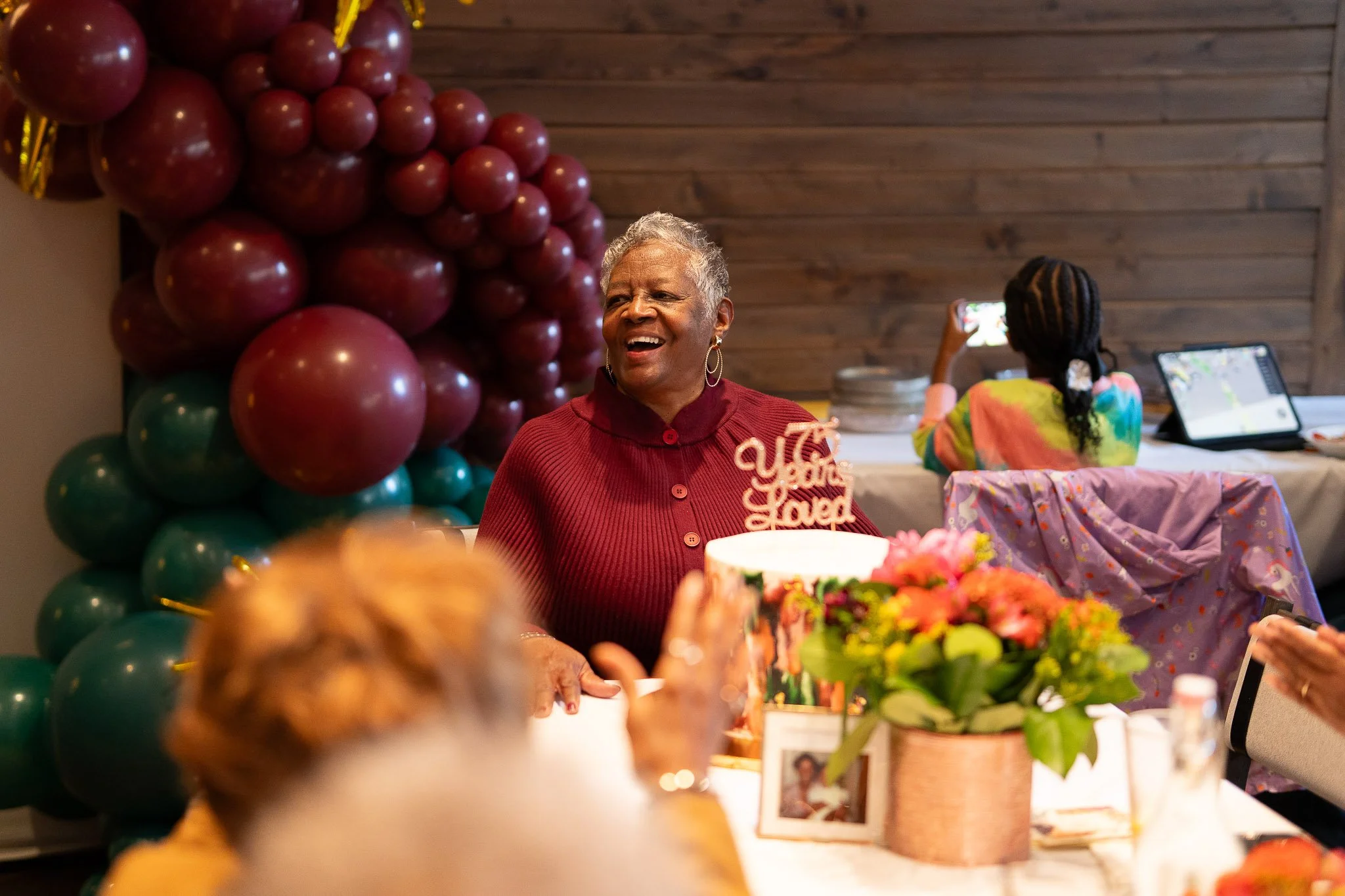 An elderly woman with short gray hair smiling at a birthday celebration, sitting at a table with flowers and a cake. Balloons are in the background, and a young girl with braided hair is taking a photo with a smartphone.