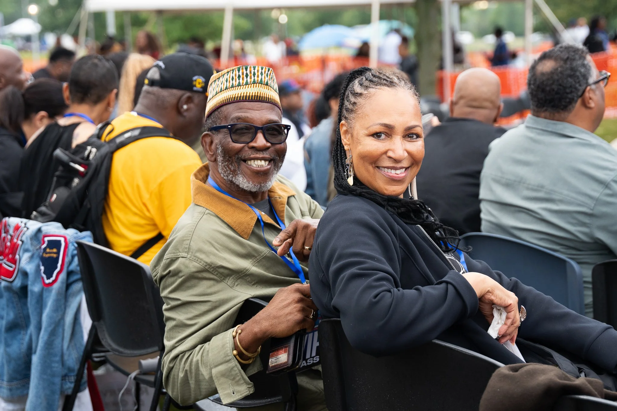 Smiling man with glasses and a woman sitting outdoors at a large event or gathering, surrounded by other attendees.