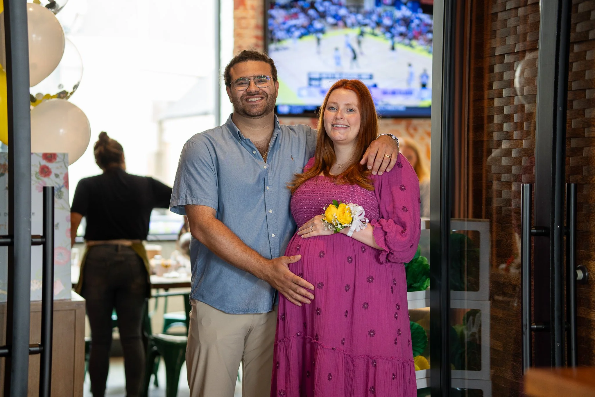 A smiling man with glasses and a woman with red hair, holding a yellow flower bouquet, stand together in a restaurant or cafe. The woman appears to be pregnant. There is a TV screen in the background and a person in black attire in the background.