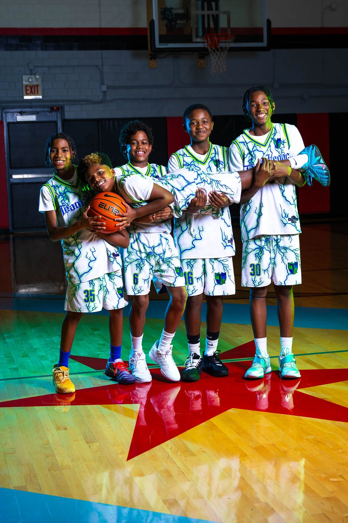 A group of five young basketball players in uniform, with one holding a basketball, standing on a gymnasium floor, smiling and posing for a photo.