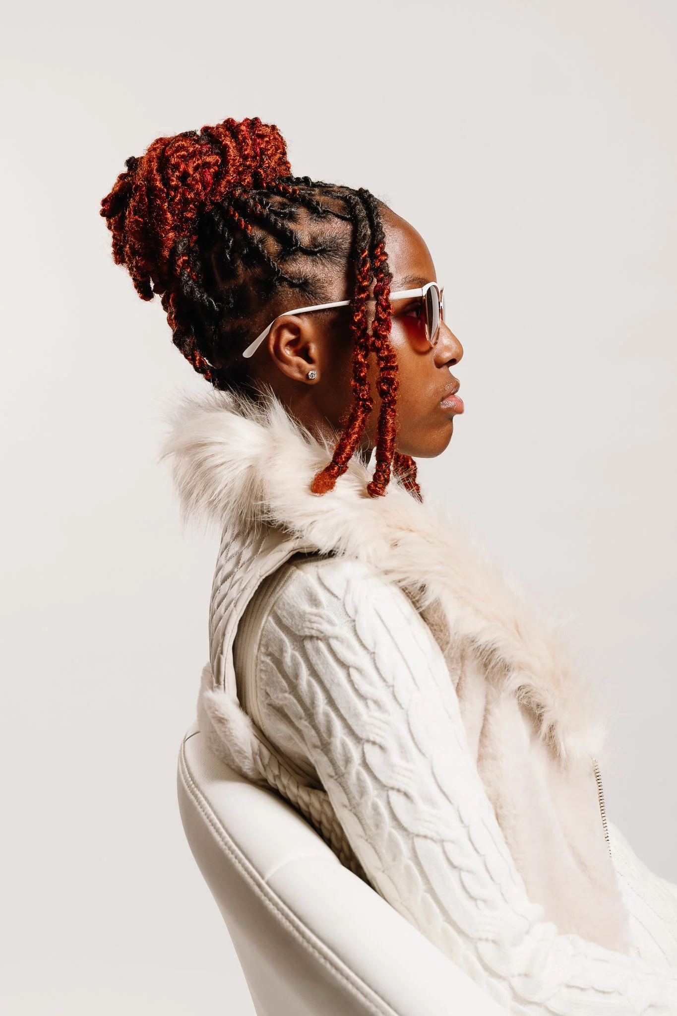 Side profile of a woman with red and black dreadlocks wearing sunglasses, a white fur-lined jacket, and earrings, sitting on a white chair against a plain background.