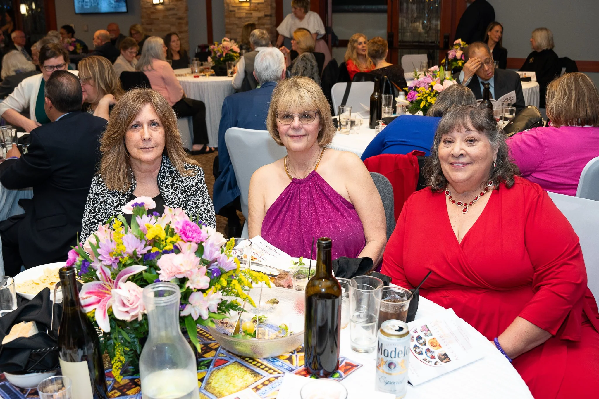 Three women sitting at a decorated banquet table with drinks and a flower centerpiece in a large, well-lit event hall filled with other guests.