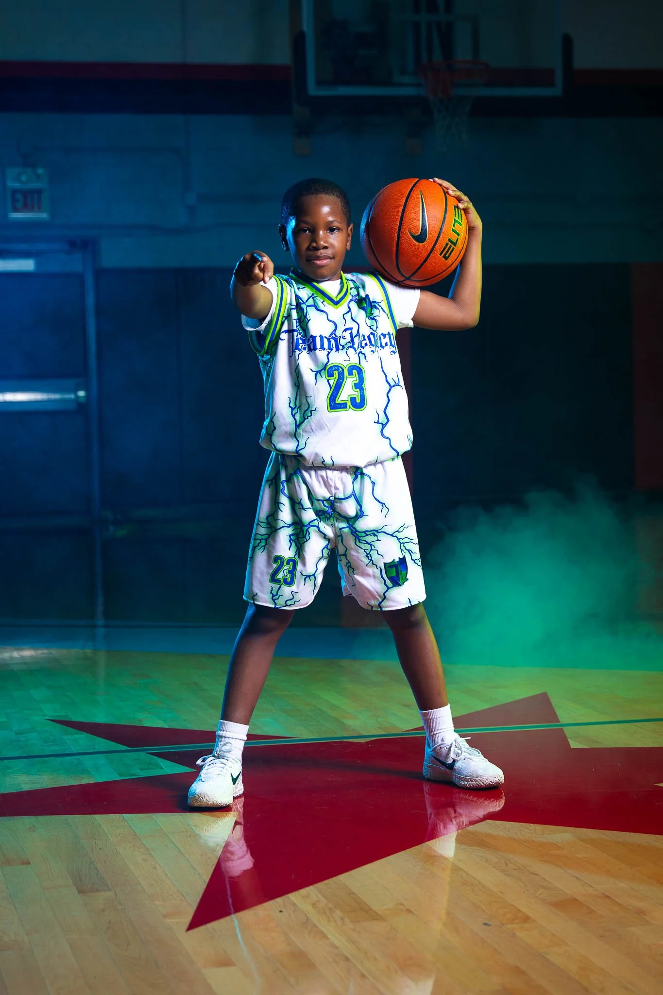 A young boy in a basketball uniform standing on a gym court, holding a basketball in one hand and pointing forward with the other. The gym has a wooden floor with a red star at the center.