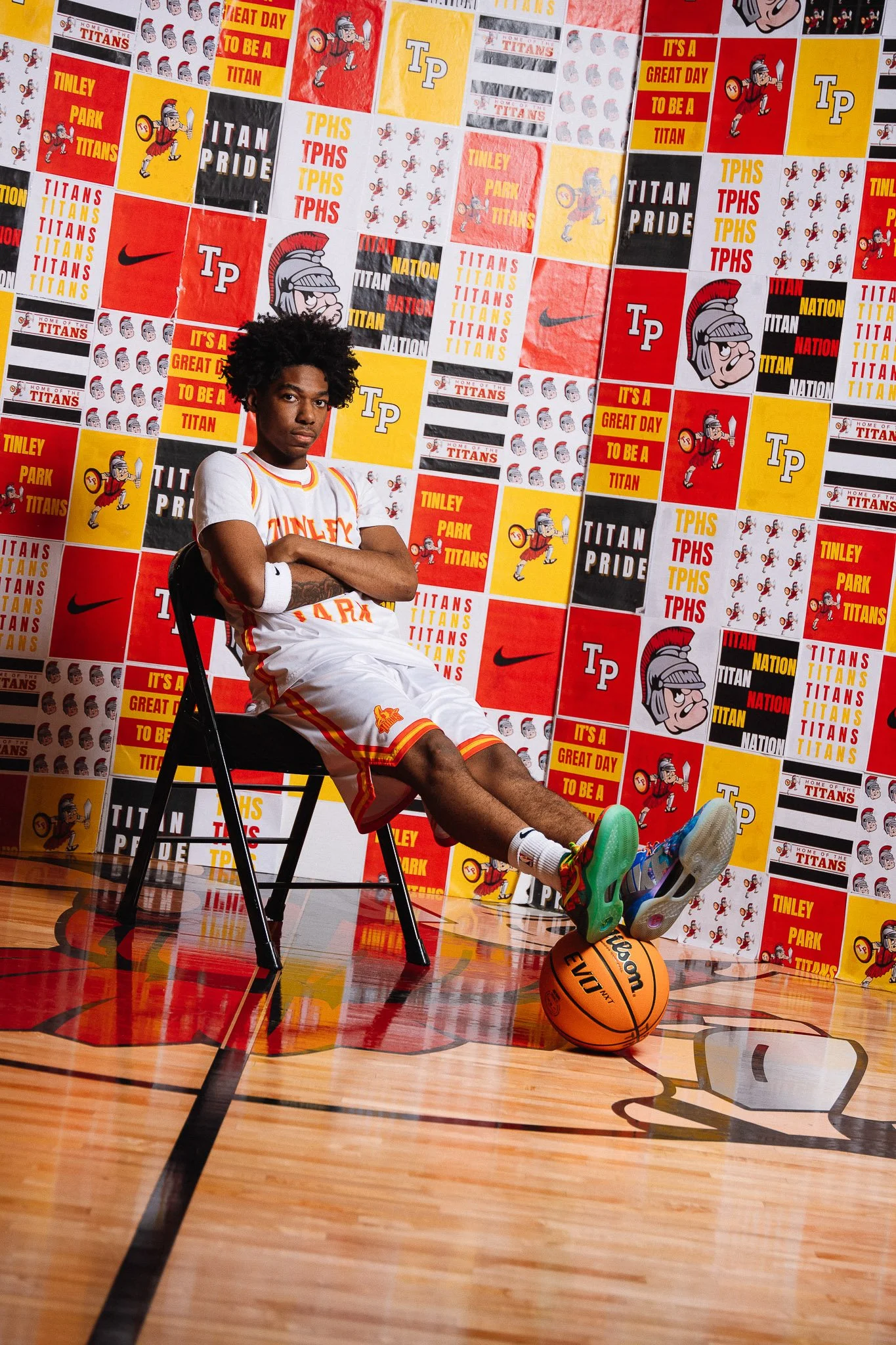 A young basketball player in a Tinley Park Titans uniform sits on a chair with arms crossed, in a gymnasium with a colorful wall covered in team and school logos and slogans. A basketball rests on the floor near his feet.