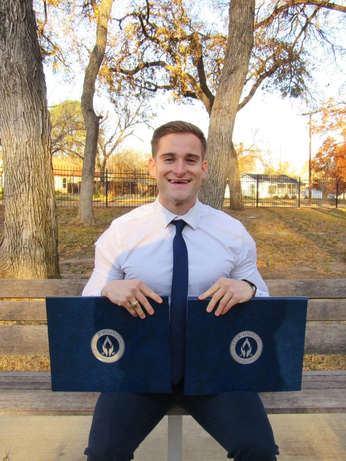 A young man with short brown hair, sitting on a park bench outdoors during autumn, holding two dark blue diploma covers with a university emblem, smiling at the camera.