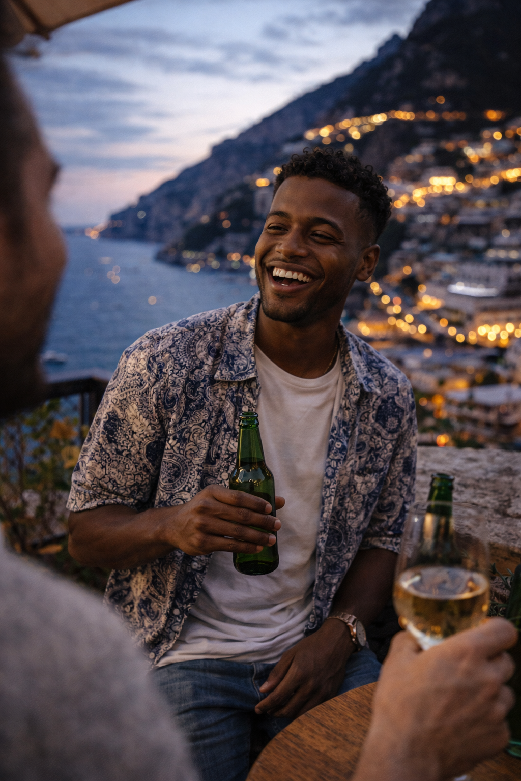 A smiling man wearing sunglasses and a beige jacket laughs while holding a coffee cup at an outdoor café.