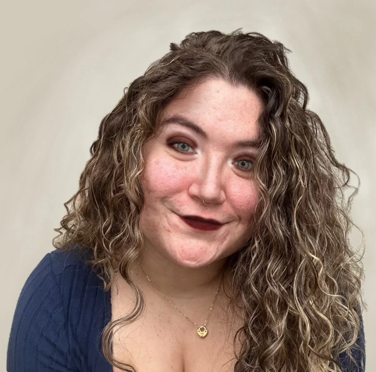 A young woman with long curly hair and blue eyes, smiling, wearing a navy top and a gold necklace with a small charm.
