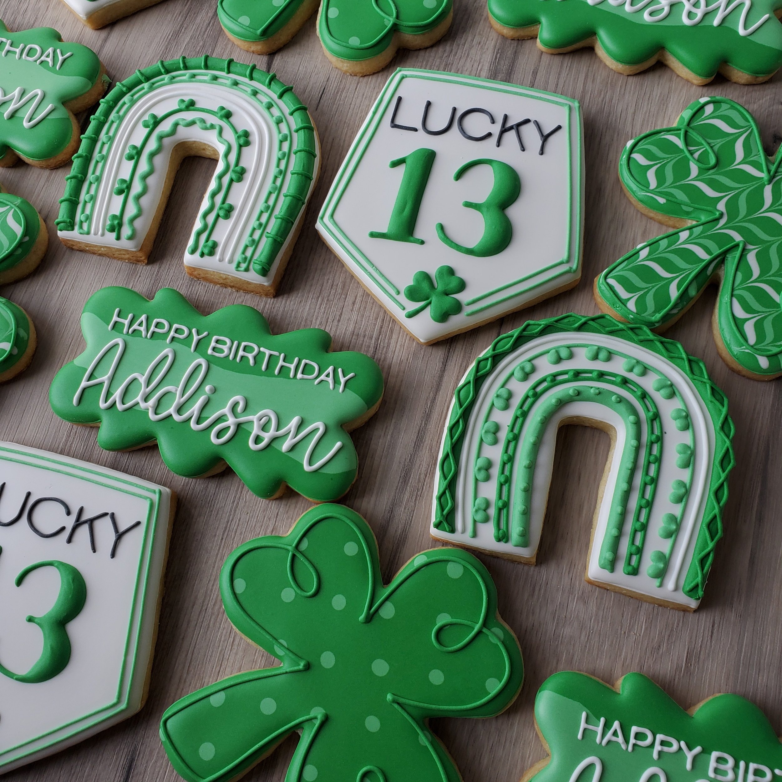 St. Patrick's Day themed decorated cookies featuring shamrocks, rainbows, a hexagon with the number 13, and birthday messages for Addison in green, white, and black icing.
