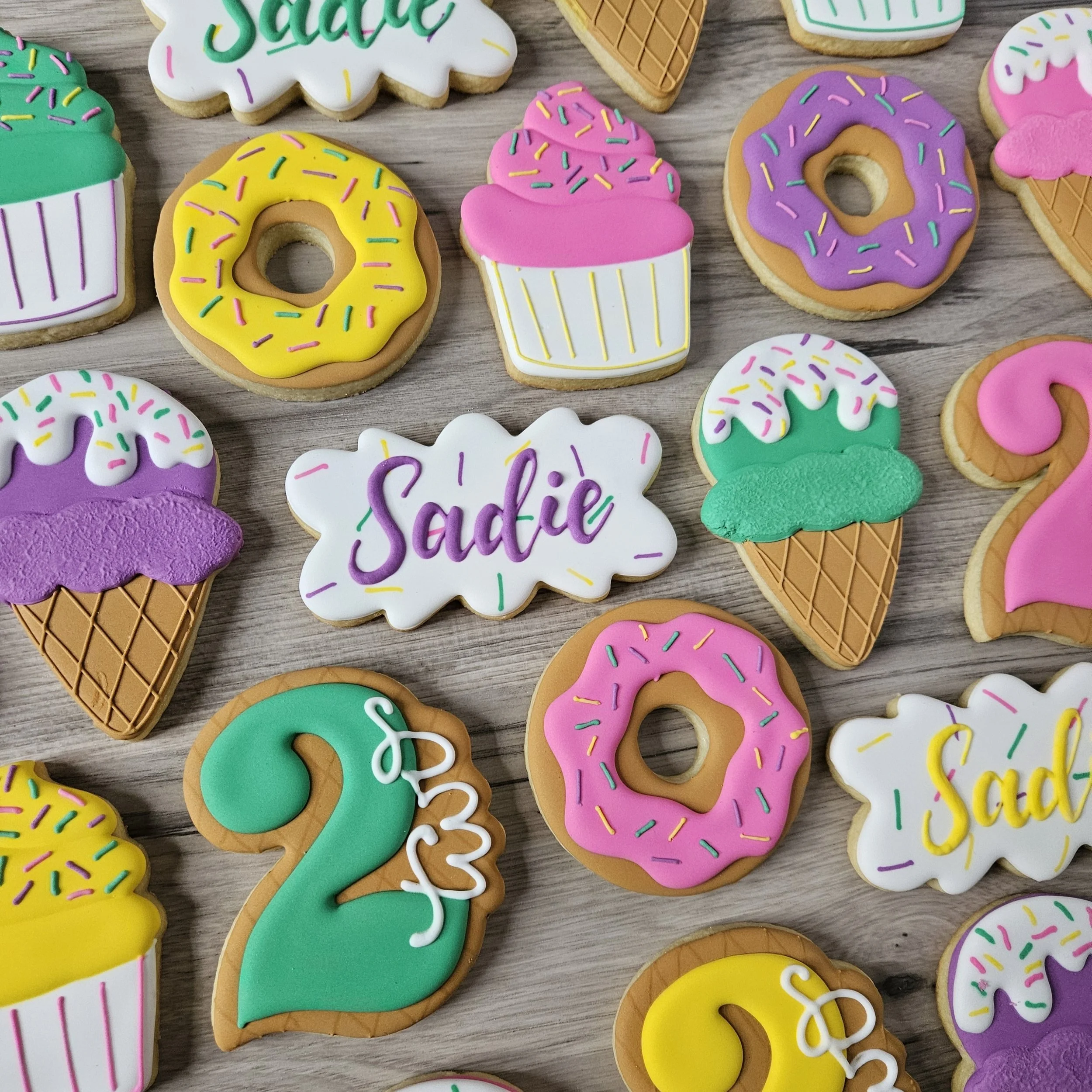 Colorful decorated cookies on a wooden surface, including cupcakes, donuts with pink, purple, and yellow icing, green ice cream cones, and cookies with the name 'Sadie' and the number '2' written on them.