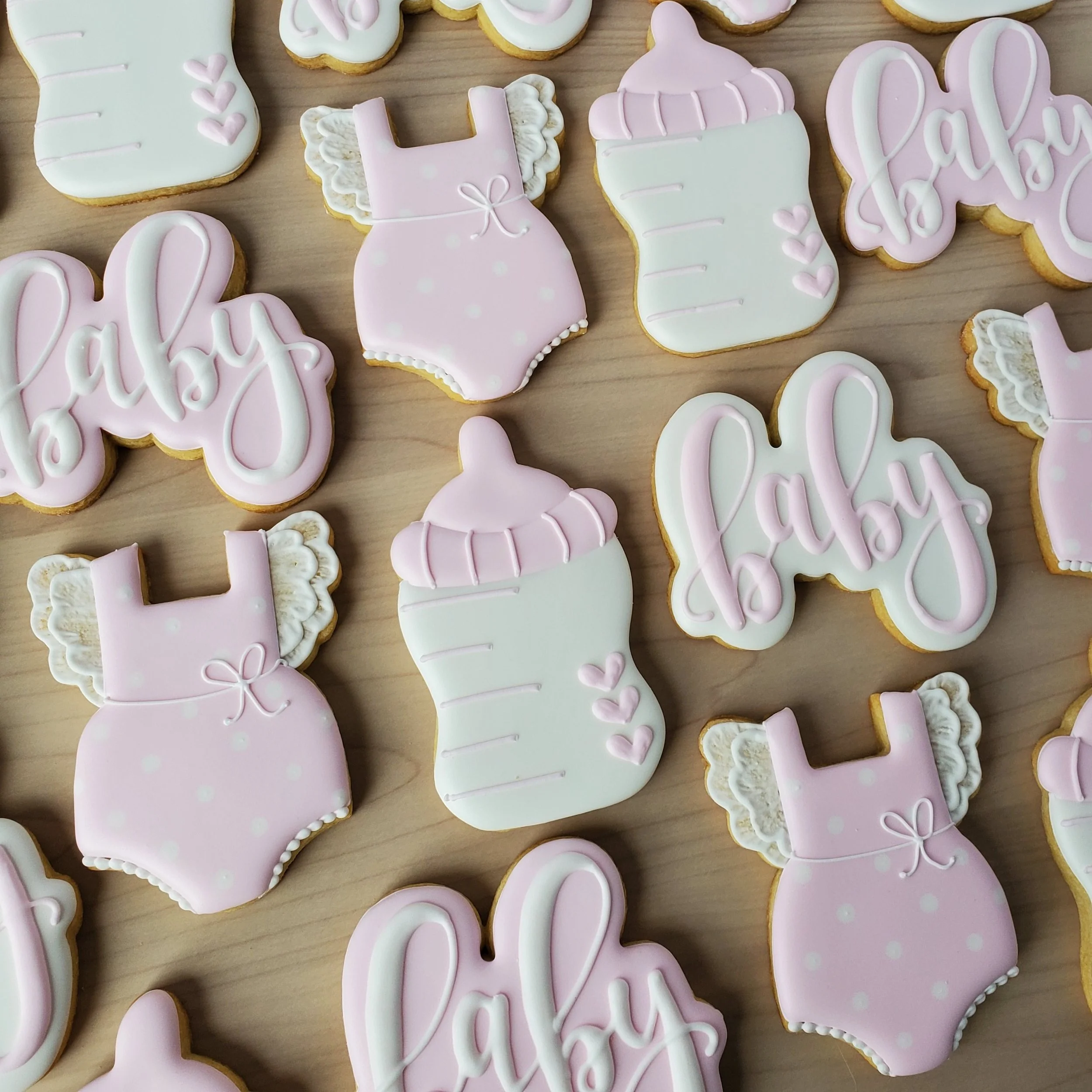 Decorated baby-themed cookies with pink and white icing, featuring words 'baby', baby bottle with pink cap and three hearts, and pink baby onesies with attached lace-like wings.