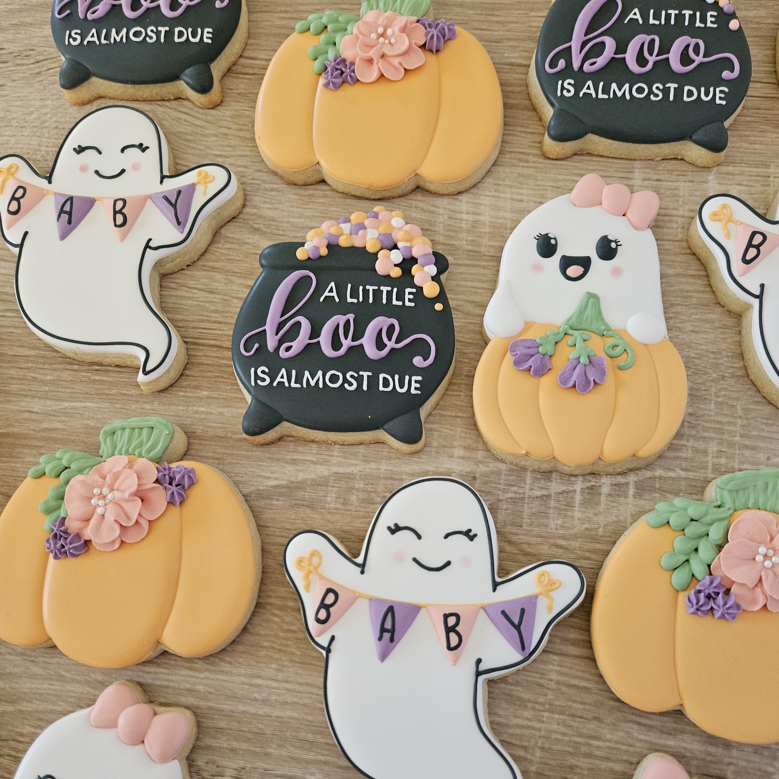Decorated Halloween-themed cookies placed on a wooden surface, featuring ghosts holding 'Baby' banners, pumpkins with flowers, and black cauldrons with the message 'A little boo is almost due'.