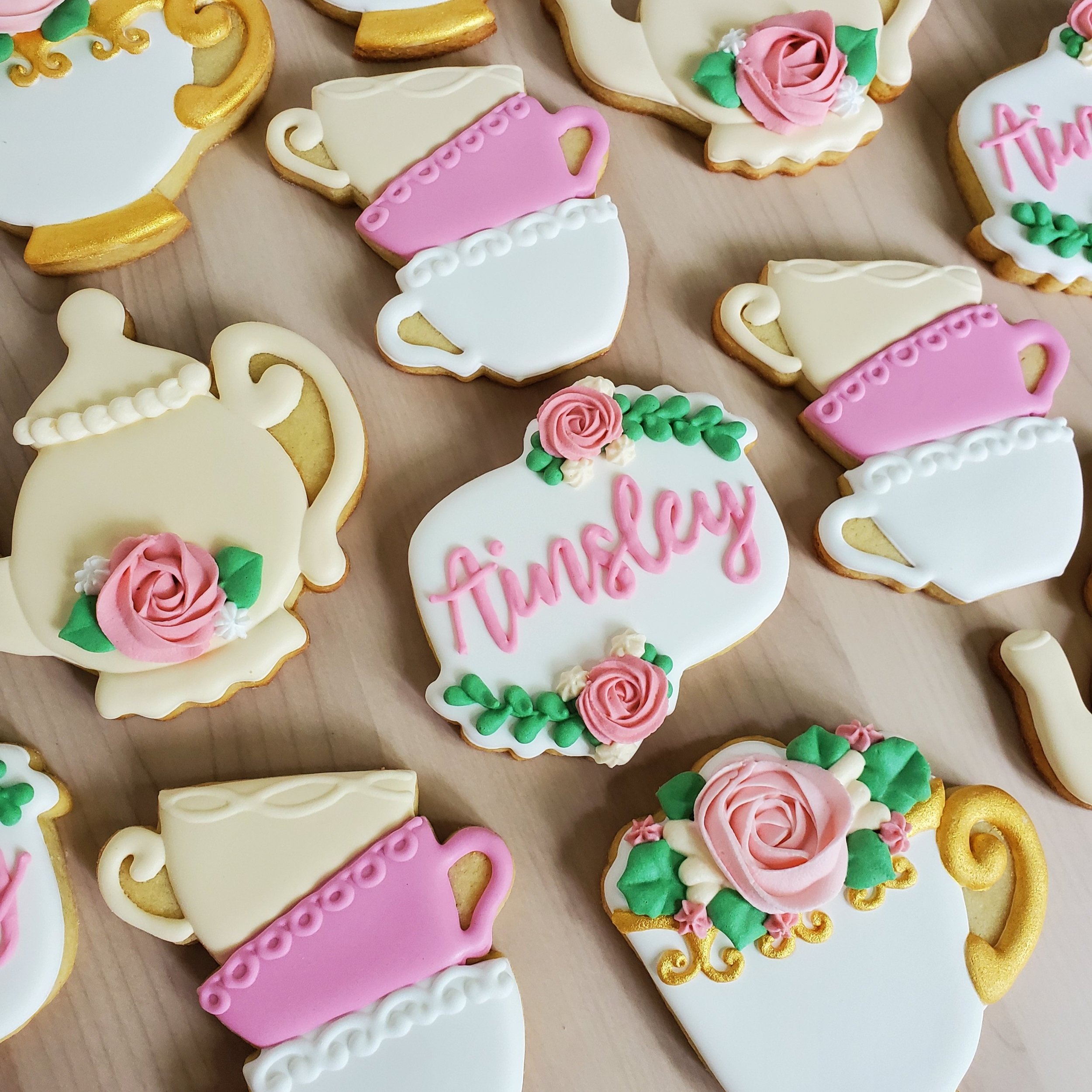 Decorative cookies shaped like teapots and teacups with floral and pink icing, one cookie with the word 'tinselry' written in pink icing, all on a wooden surface.