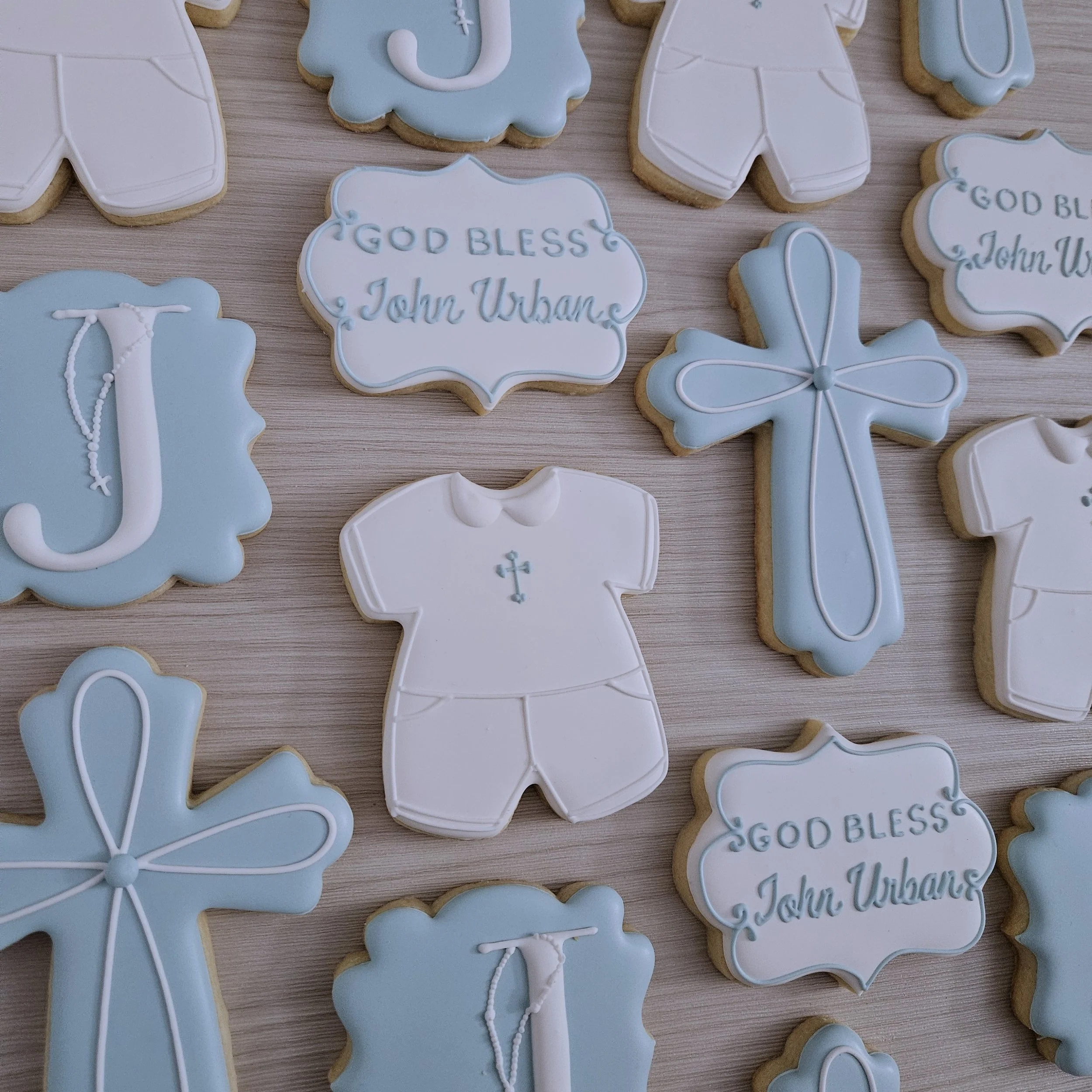 Decorative cookies with religious and baptism motifs, including a cross, a baby outfit, a Bible verse, and religious symbols in blue and white icing.