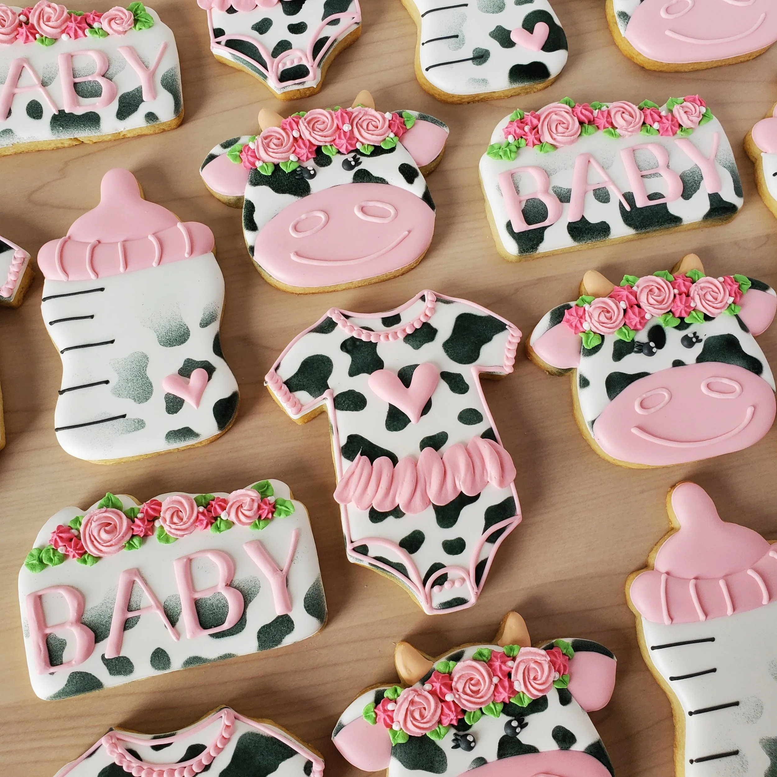 Decorated cookies with baby cow theme, including cows with pink flower crowns, a milk bottle, baby clothes, and blocks spelling 'BABY' in pink and black cow print.