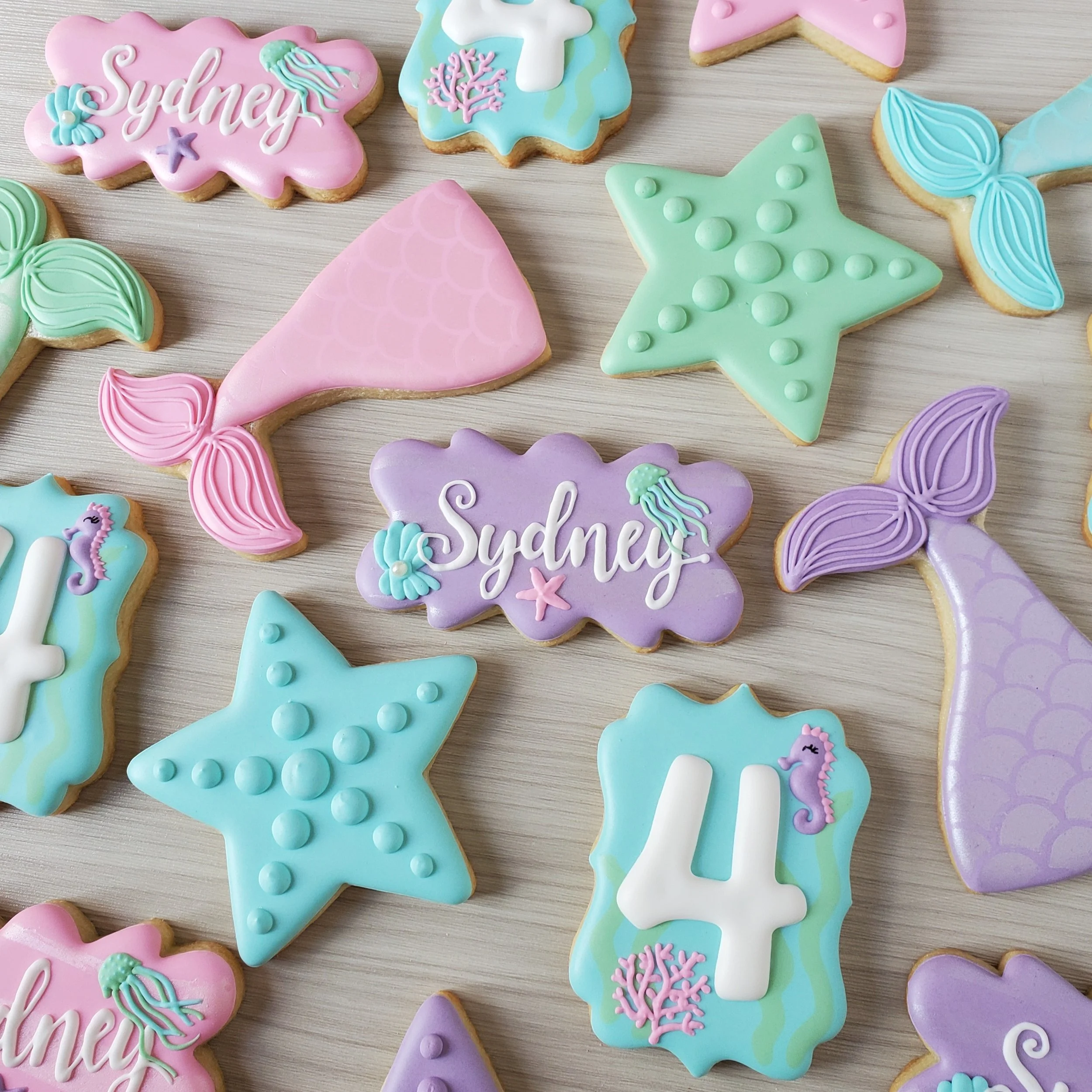 Decorated sugar cookies with a mermaid and ocean theme, featuring starfish, jellyfish, and seahorses. Some cookies have the name 'Sydney' written on them, and others have the number 4. The cookies are in pastel colors like pink, purple, blue, green, 
