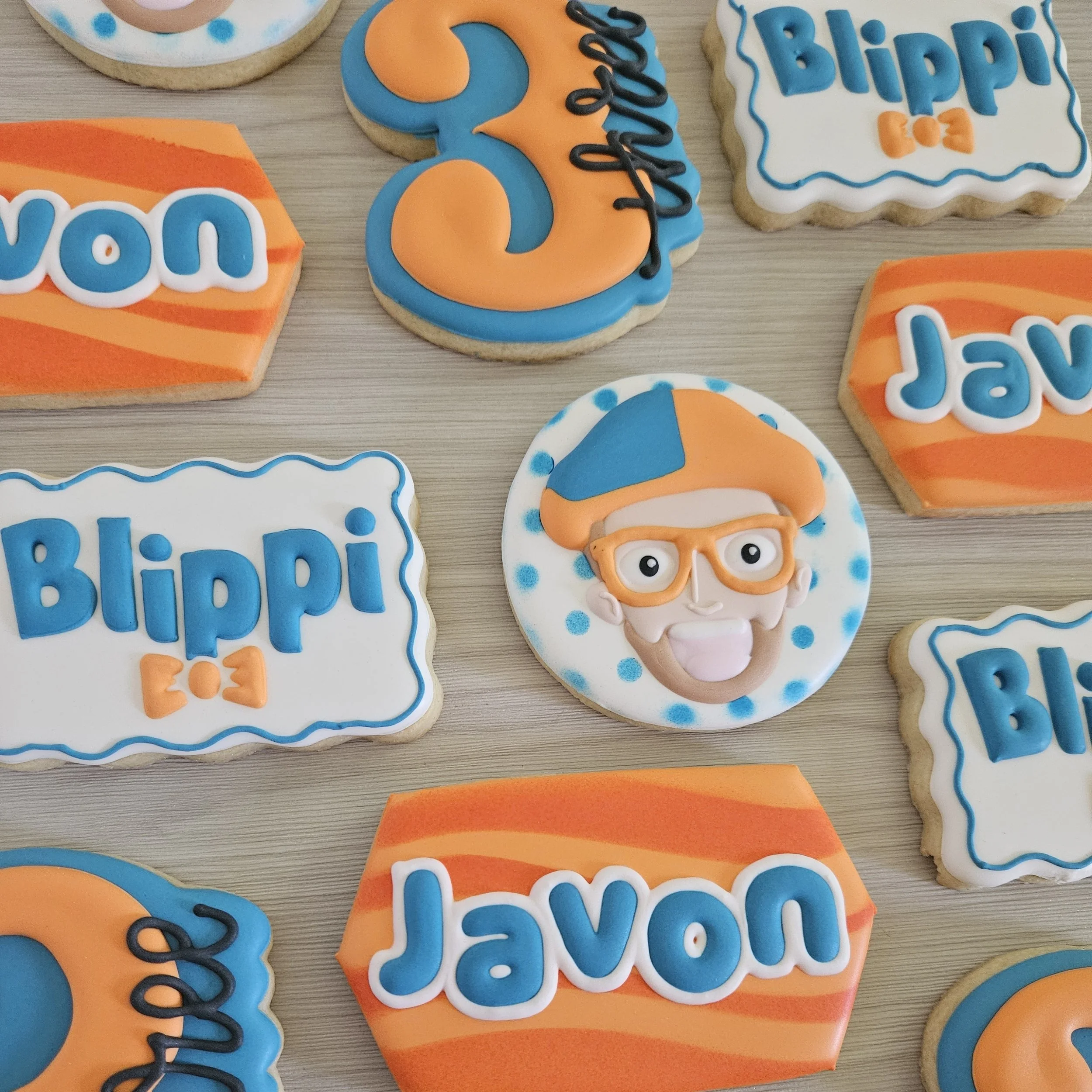 Collection of decorated cookies for a birthday celebration featuring the name Jaxon, with themed cookies including a smiling man with glasses and a baseball cap, and cookies with the name 'Blippi' and playful orange and blue designs.