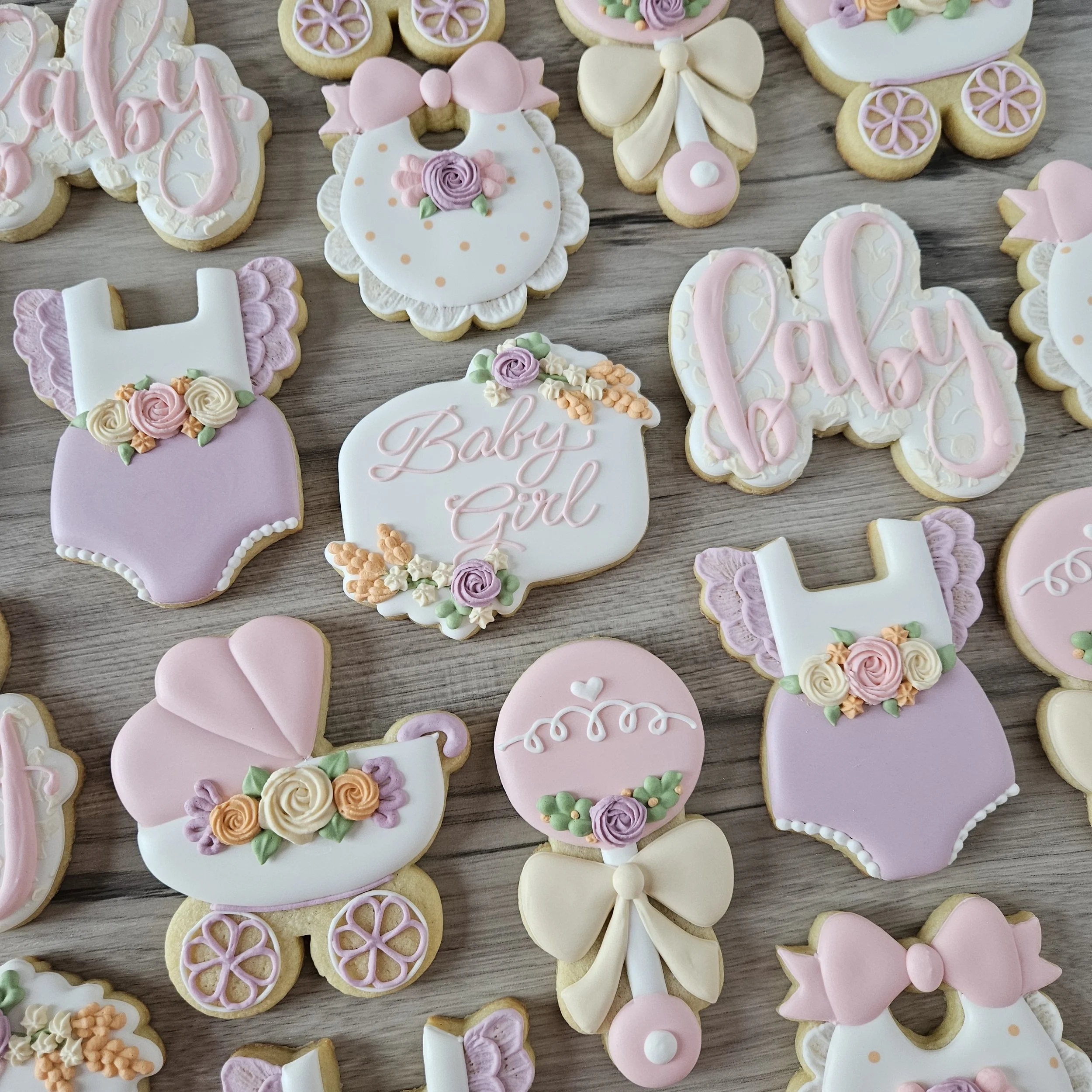 Decorated cookies for a baby shower, featuring pastel pink, purple, yellow, and white colors with baby-themed designs including bottles, strollers, bows, and the words "Baby Girl" and "baby".