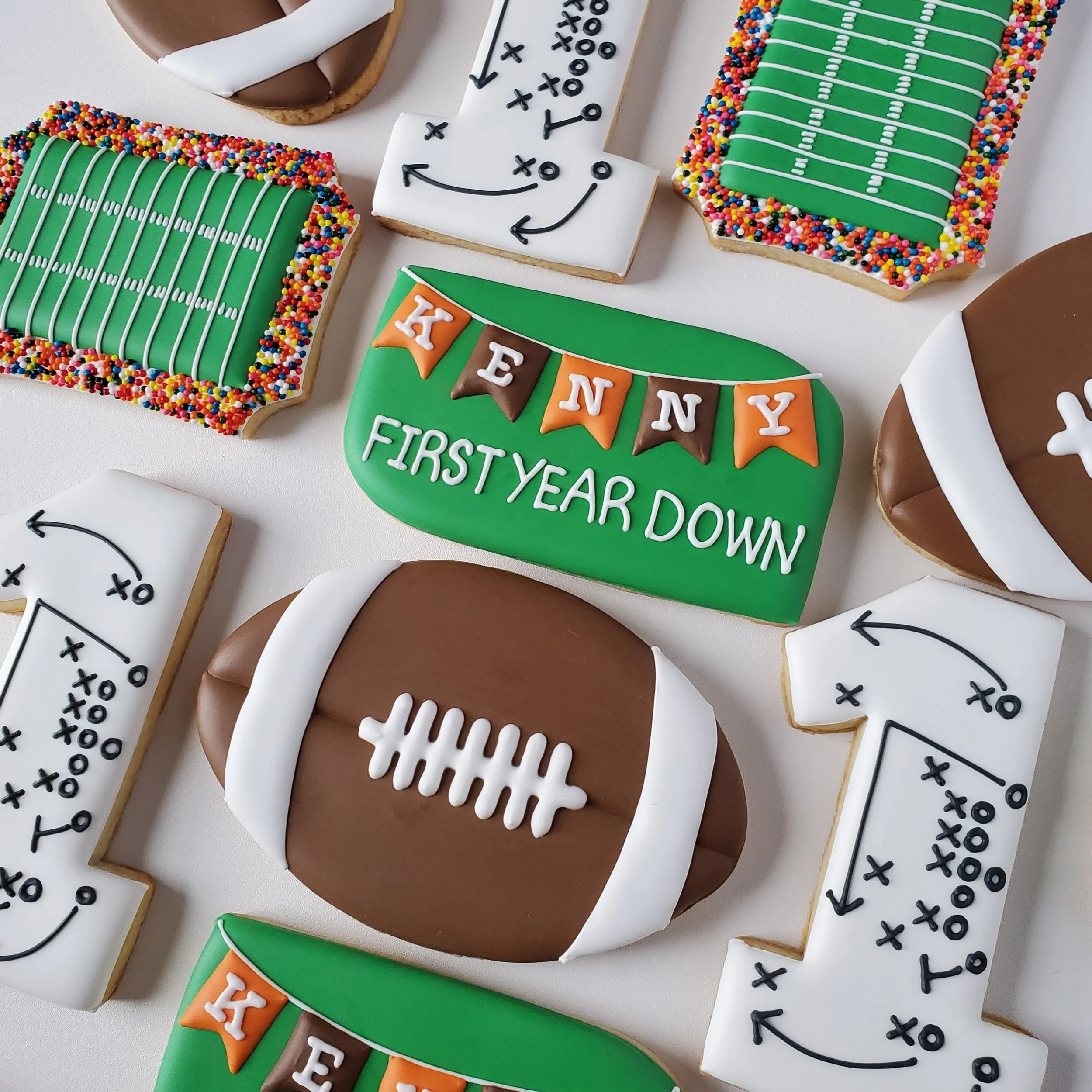 Assorted decorated cookies for a First Down themed first birthday with football shapes, a banner reading "Kenny first year down", a number one with a x-o football play on it, and a football stadium with colorful sprinkles and field markings