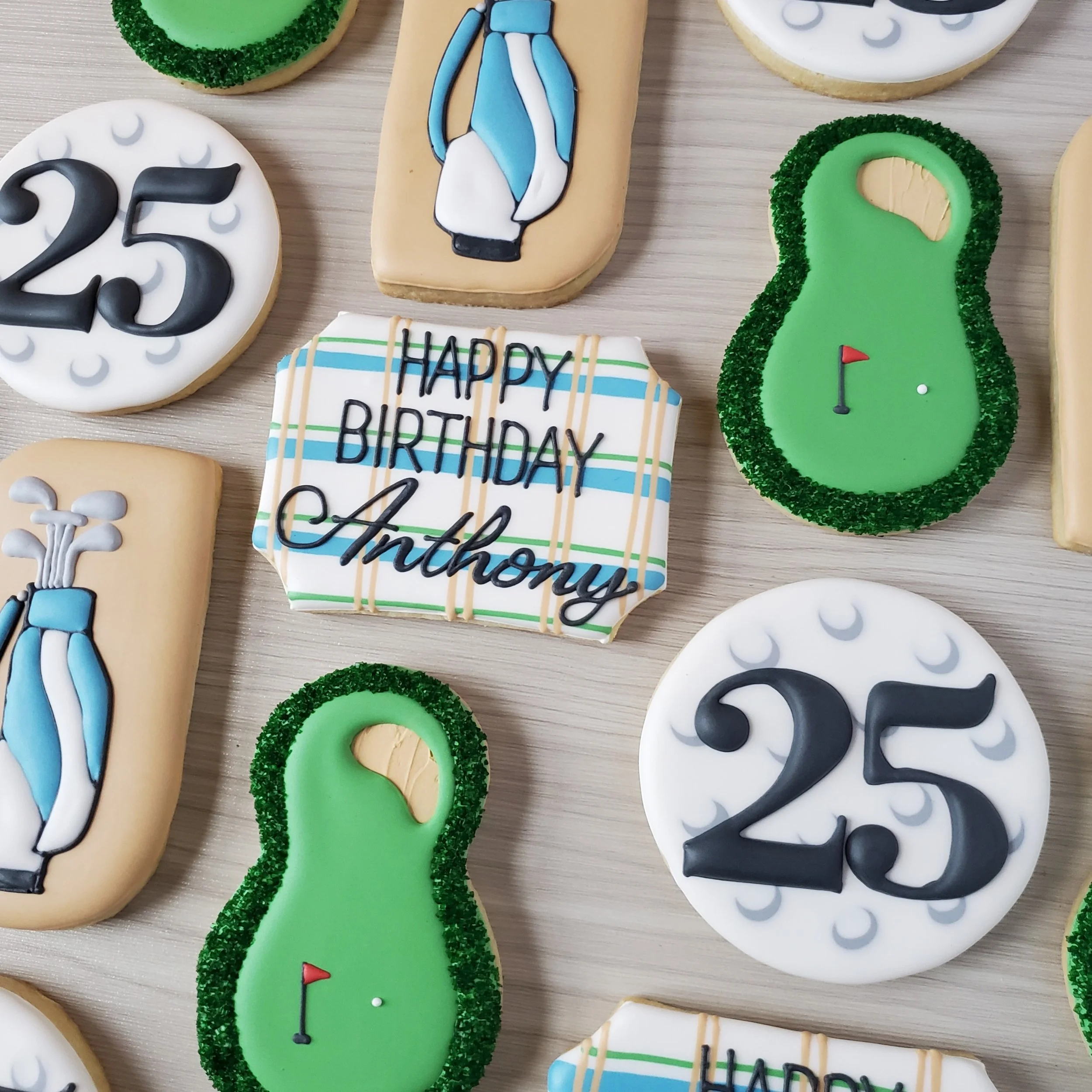 Assorted birthday cookies decorated with golf themes, including golf balls, golf clubs, a golf bag, and a golf course flag. One cookie has a birthday message in black and blue icing, reading 'HAPPY BIRTHDAY Anthony,' with a plaid background. The cook