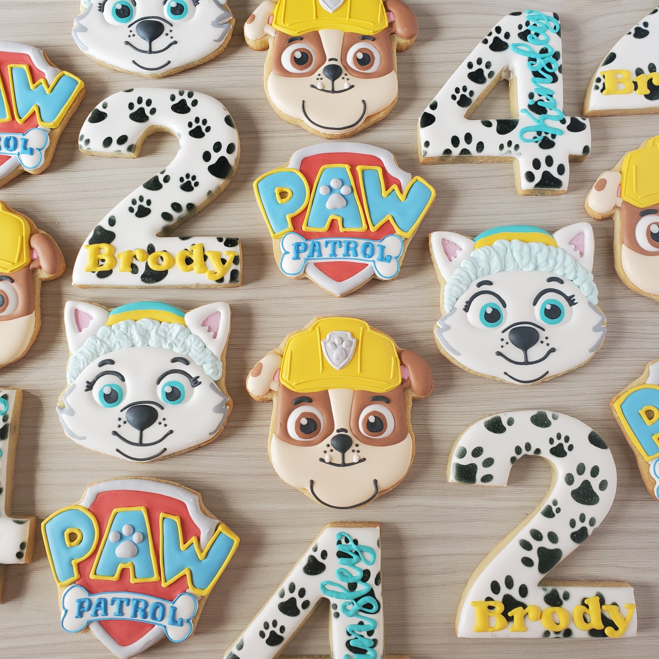 Decorative cookies themed with PAW Patrol characters and numbers including a husky, and bulldog with construction helmets, and cookies shaped as the number 2 and 4 with paw print patterns, all on a light wooden surface.