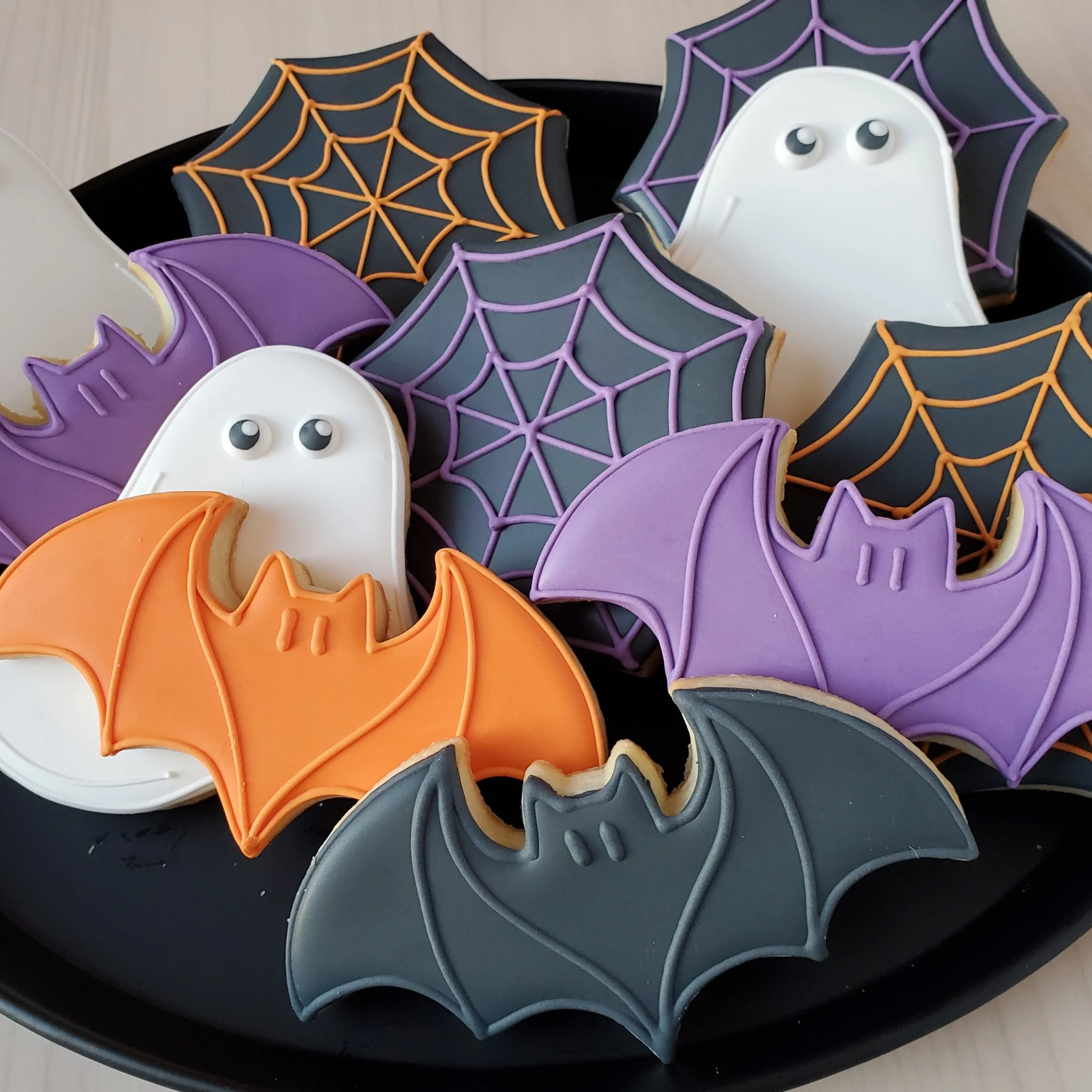 Halloween cookies decorated as ghosts, bats, spider webs, and bats with icing in orange, purple, black, white, and gray colors, arranged on a black tray.