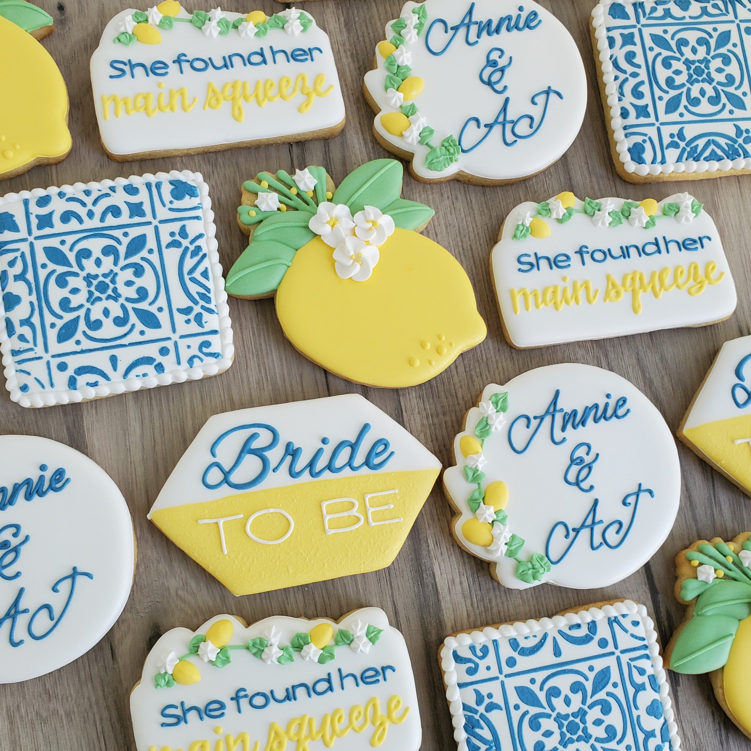 Decorated cookies for a bridal shower with blue, white, and yellow icing. Some cookies have phrases like 'She found her main squeeze,' 'Bride to be,' and 'Annie & A.' Others feature lemon and floral designs, as well as intricate blue and white patter
