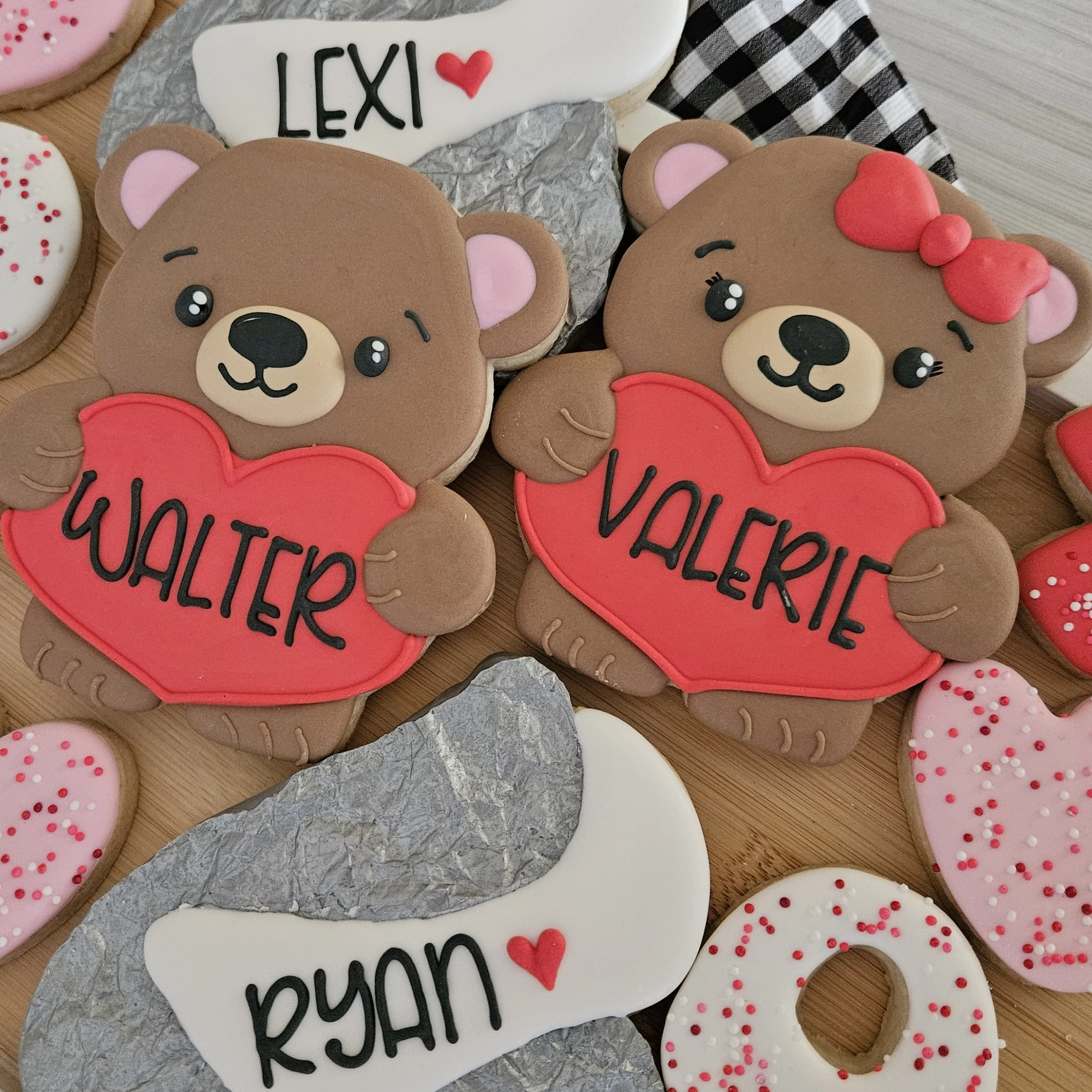 Decorative cookies shaped like teddy bears holding red hearts with names Walter and Valerie, surrounded by various other decorated cookies, including hearts and a bone-shaped cookie with the name Ryan.