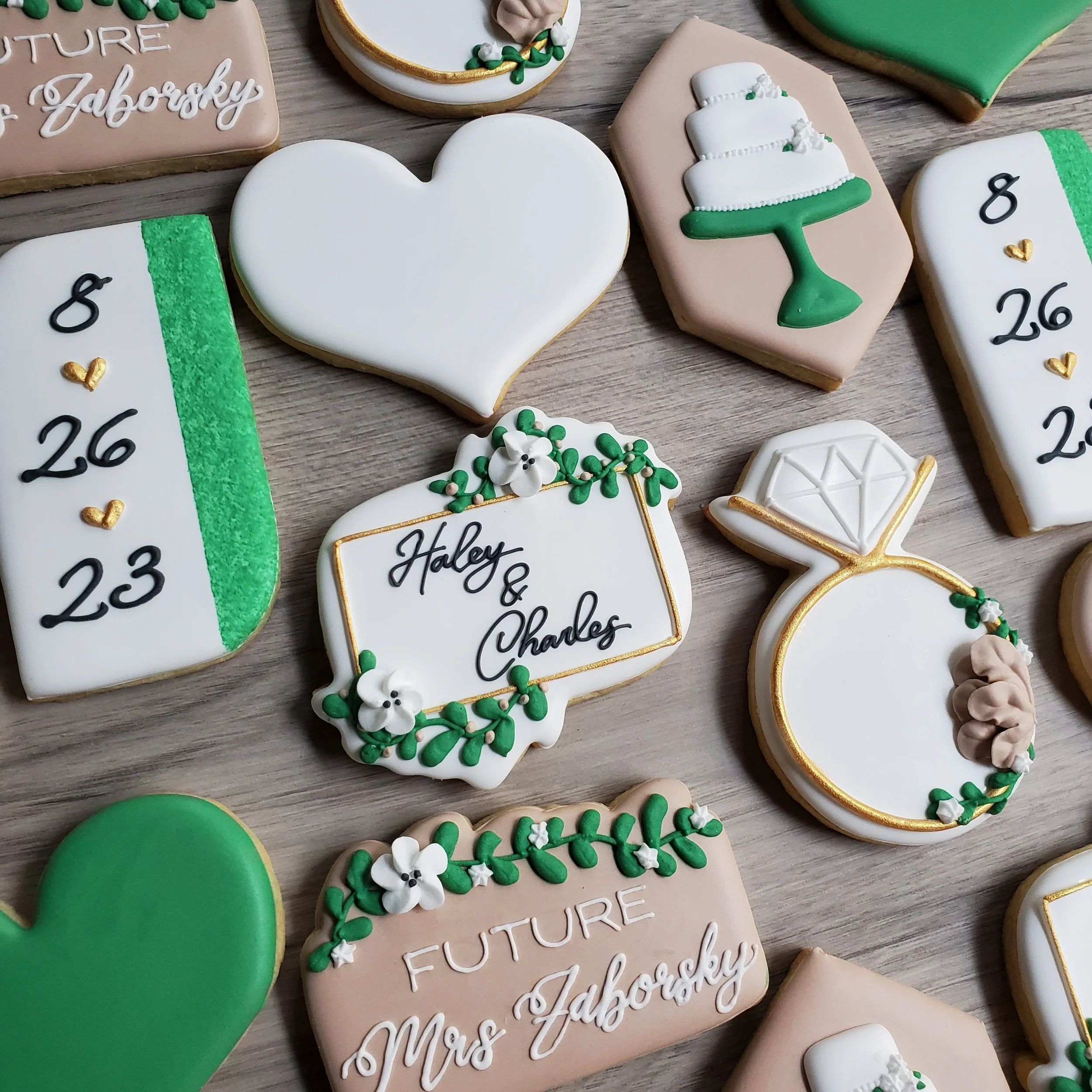 Decorated cookies for a wedding or celebration, including hearts, a diamond ring, a cake on a stand, calendars with dates, and a plaque with a wedding name. The cookies are decorated with white, green, beige, and gold icing.