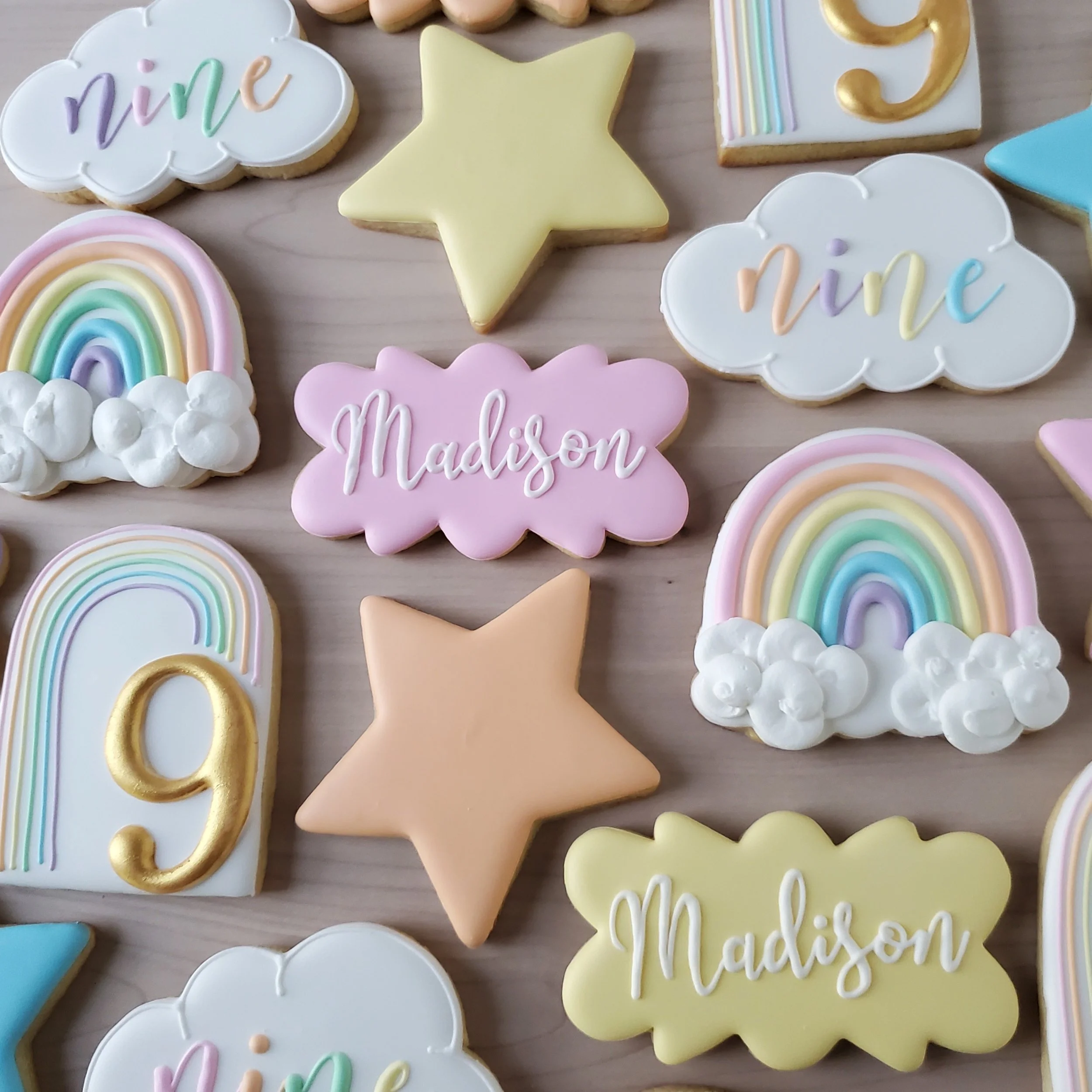 Decorative cookies in various pastel colors with rainbow, cloud, star, and number designs, some featuring the names Madison and nine.