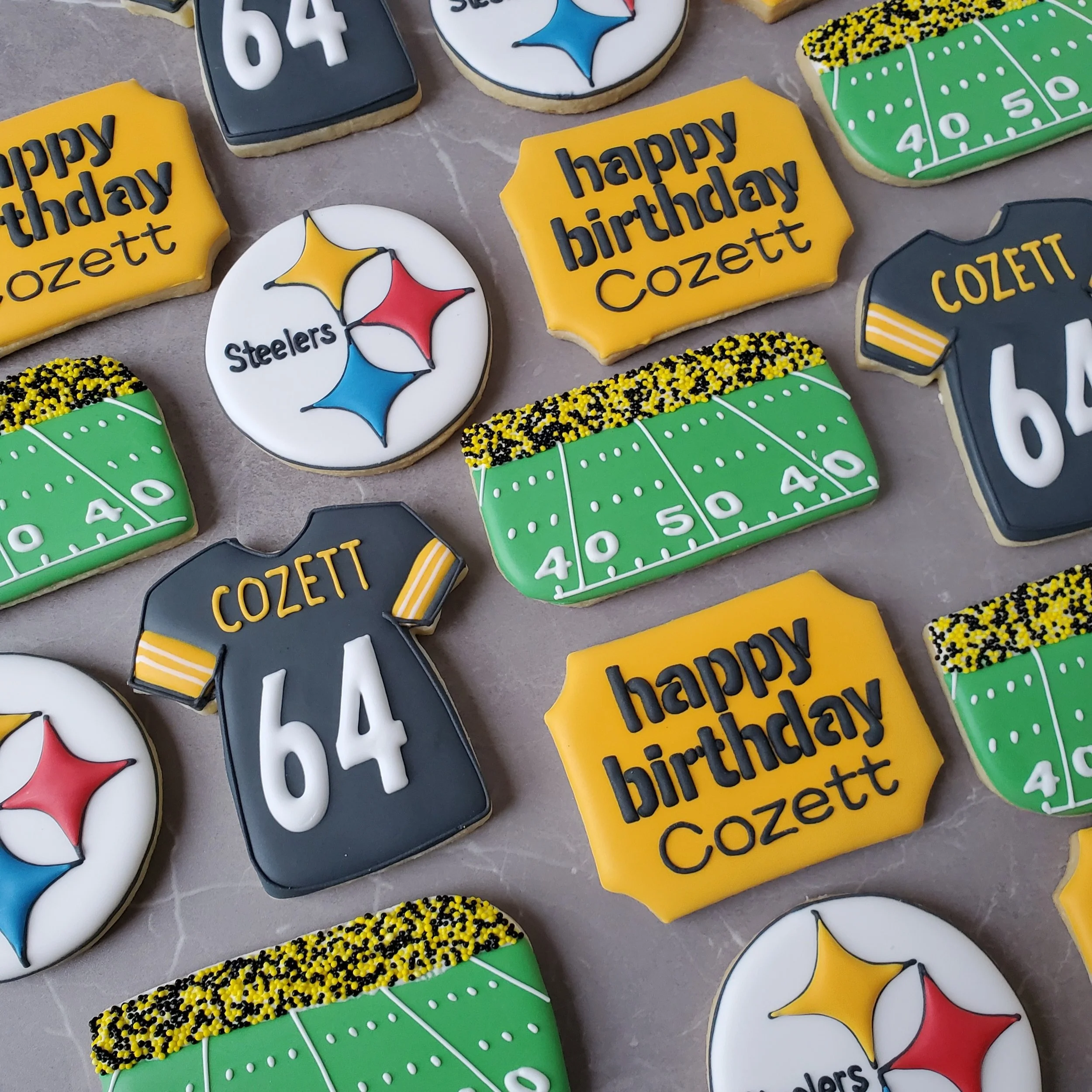 Decorated cookies celebrating a Pittsburgh Steelers themed birthday, featuring football field designs, a football jersey with the number 64 and the name Cozett, and the Steelers logo.
