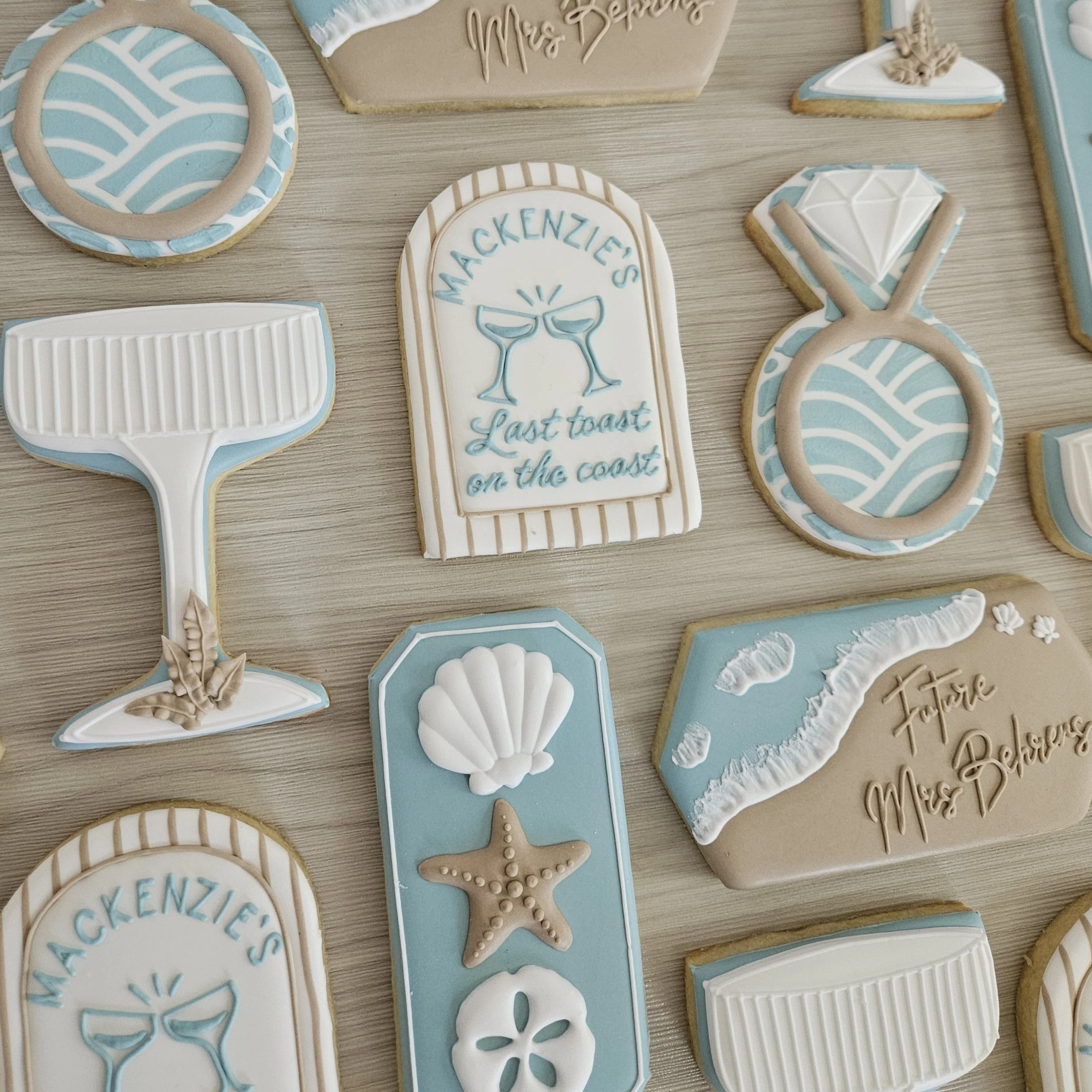 Assorted beach-themed decorated cookies in pastel blue, white, and beige colors featuring designs like seashells, starfish, cocktails, a diving mask, and messages such as "Mackenzie's Last Toast on the Coast" and "Future Mrs. Beaches".