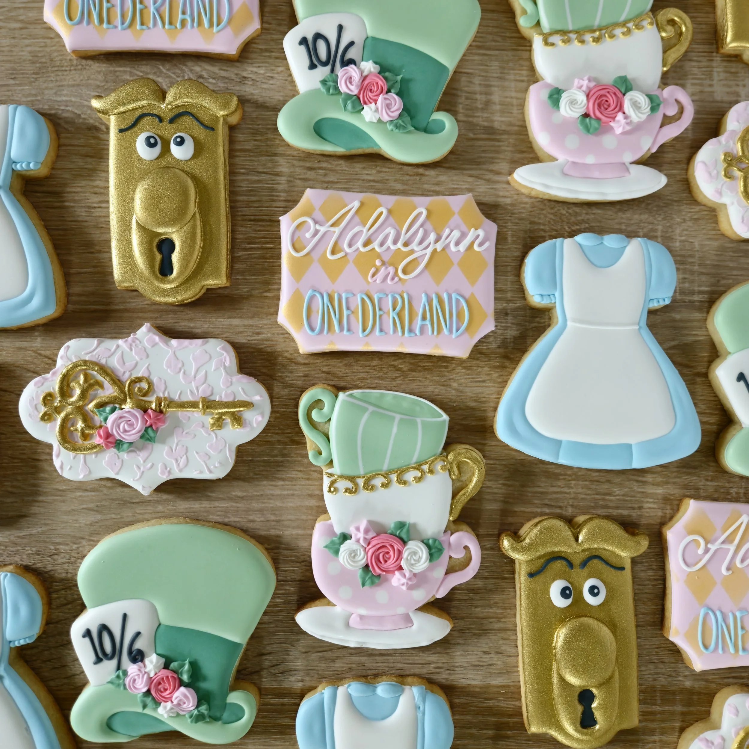 Decorated cookies themed from Alice in Wonderland, including a golden door knob, a green top hat with a white card labeled '10/6' and pink roses, a pink teacup with roses and gold accents, a blue and white dress, and a sign that reads 'Adalynn in Won