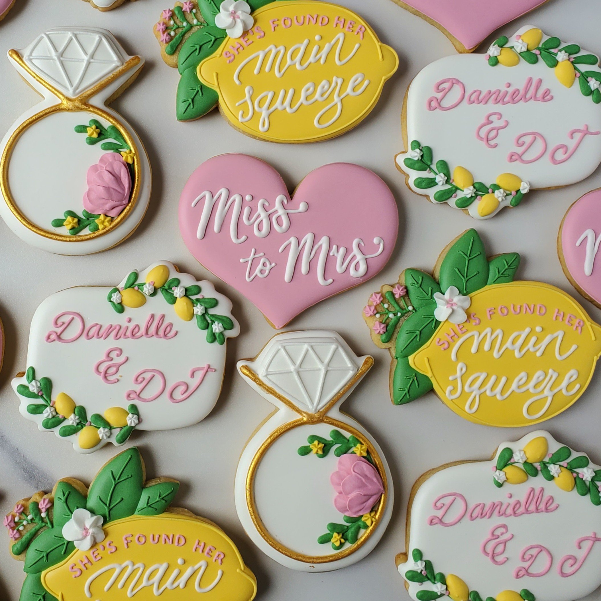 Decorated sugar cookies with various designs and phrases for a wedding. Some cookies are shaped like diamonds, hearts, and oval plaques. The cookies feature pink, yellow, white, and green icing, with phrases such as "She's Found Her Main Squeeze," "M